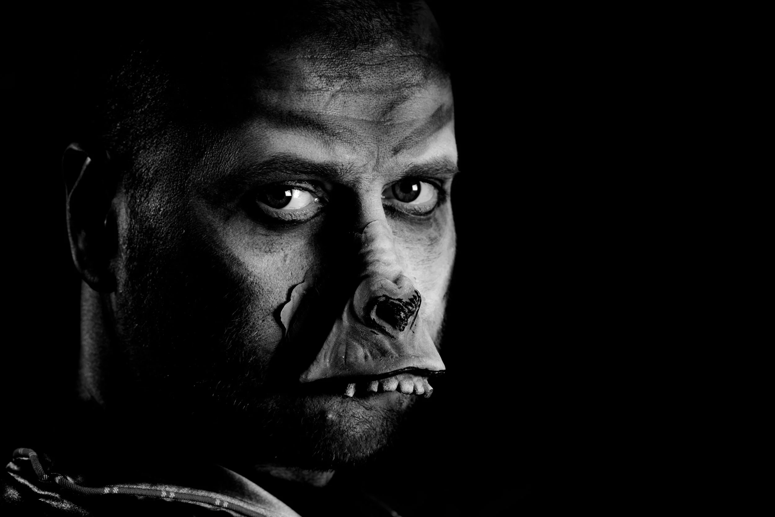 Black and white photo of a man's face with a painted clown mask covering the lower half, intense gaze, dark background.