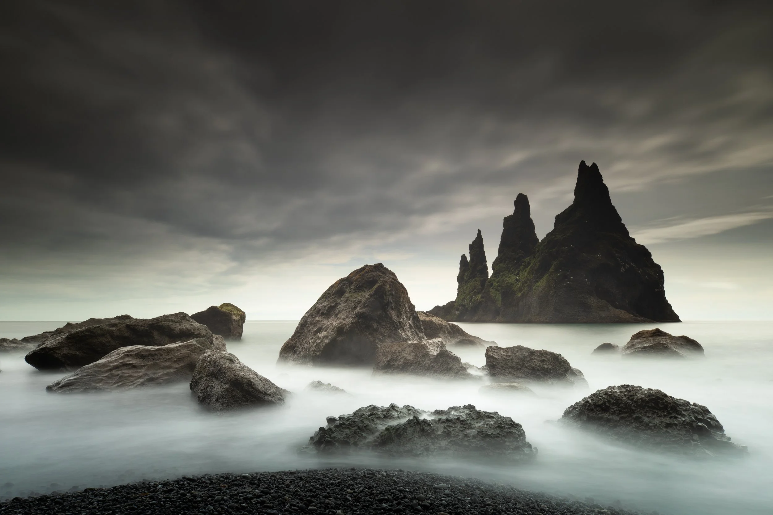 Dark stormy sky over large rock formations emerging from a foggy ocean, with smaller rocks in the foreground.