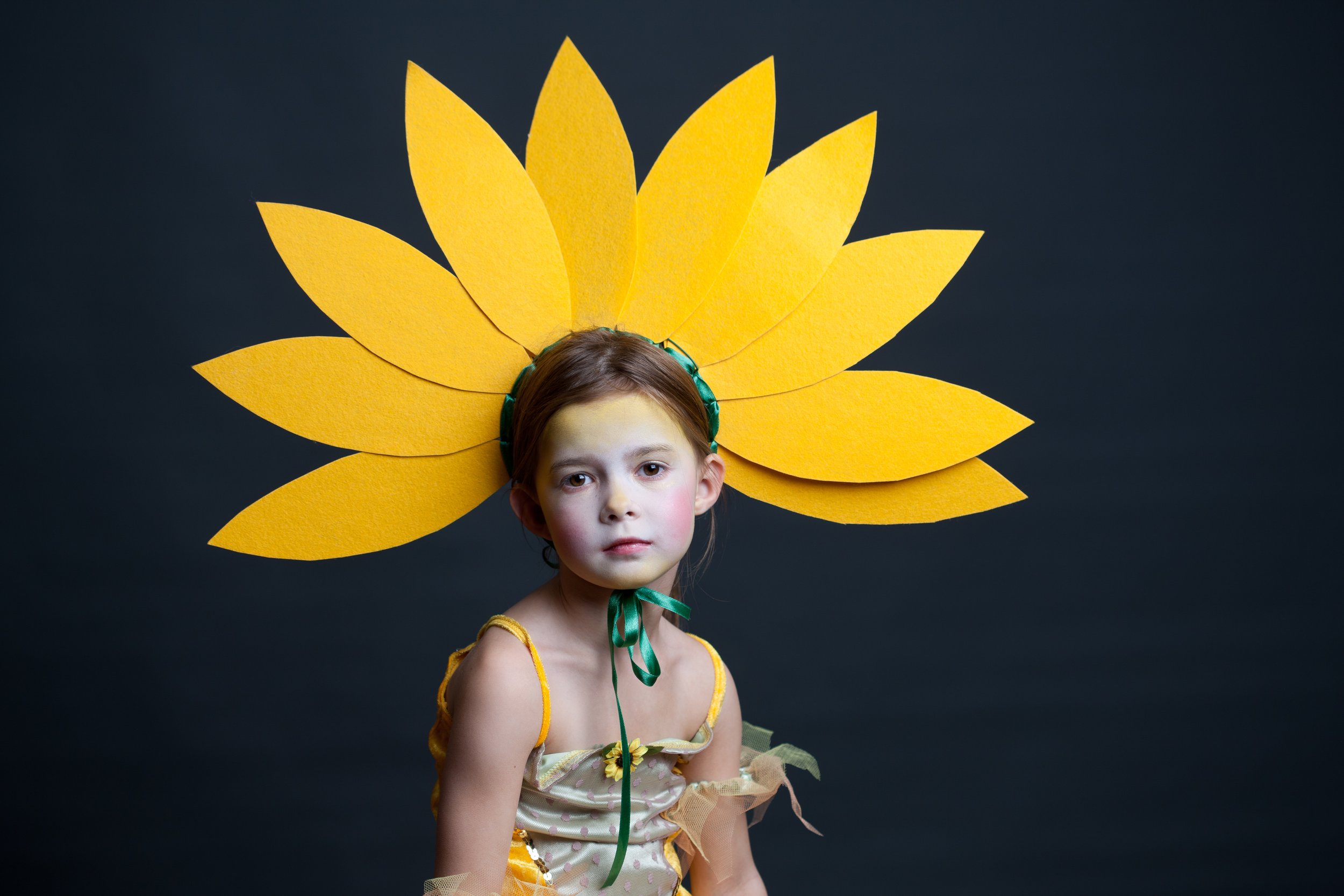 A young girl dressed as a sunflower, with a yellow petal headpiece and a yellow dress, poses against a dark background.