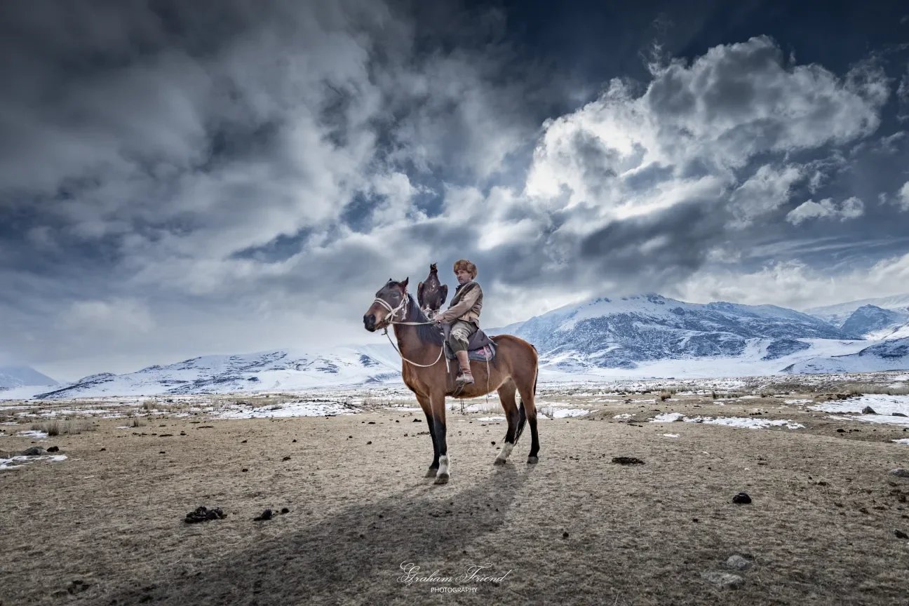 A person riding a horse in a rugged, snow-covered landscape with distant mountains and a cloudy sky.