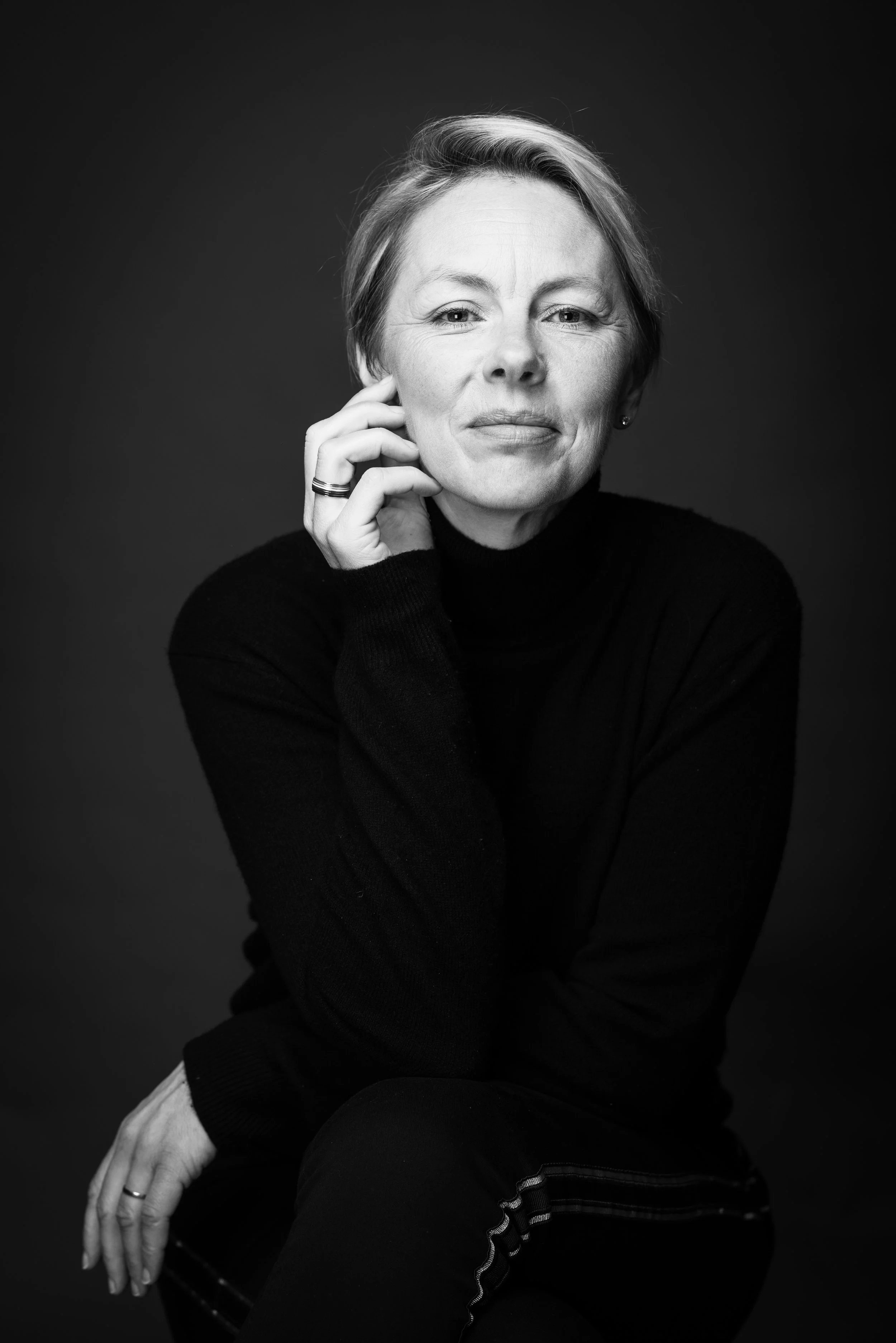 Black and white portrait of a woman with short hair, wearing a turtleneck sweater, smiling gently, sitting with her hand near her face on a dark background.