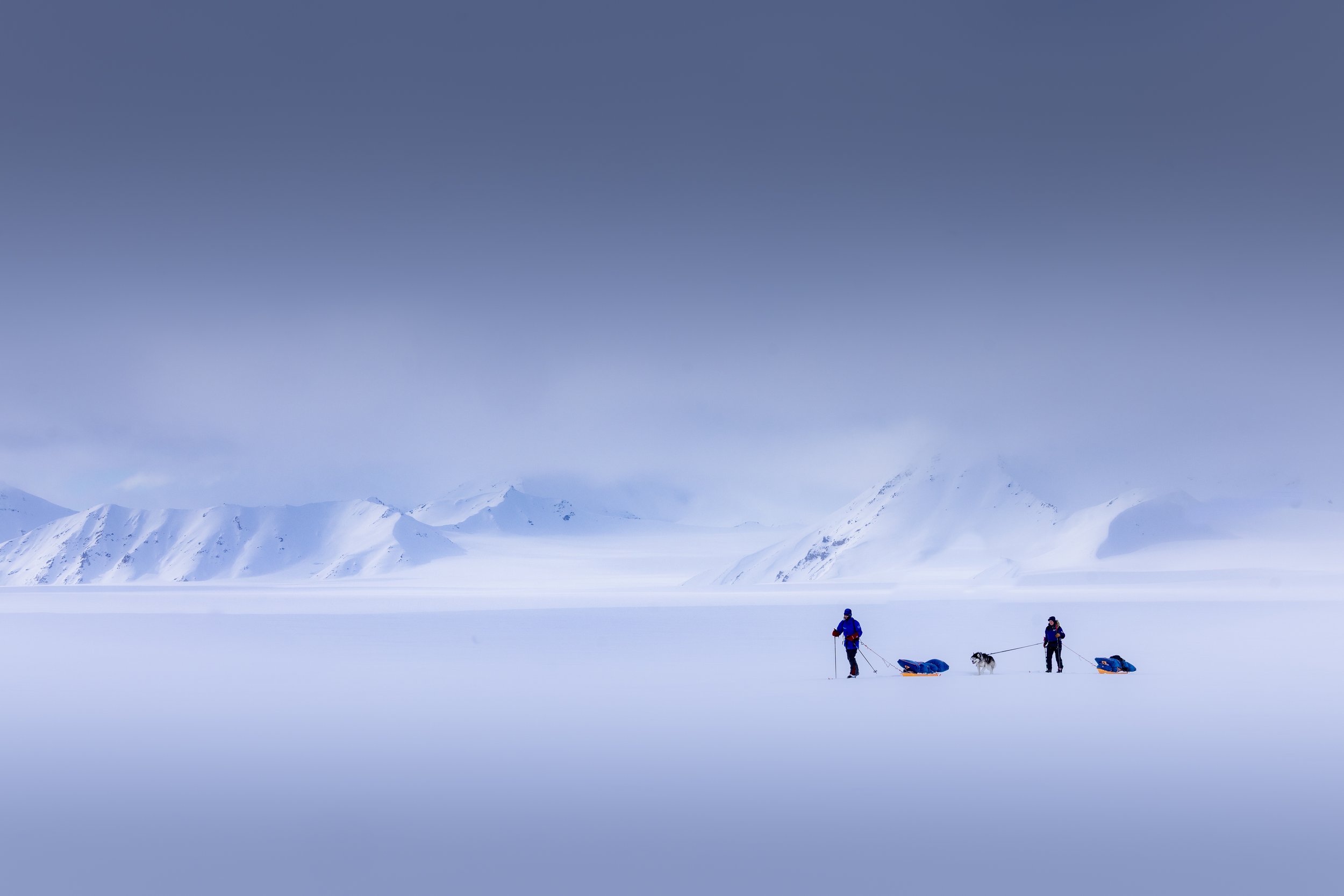Two explorers in cold-weather gear pulling sleds across a snow-covered landscape with snow-capped mountains in the distance and a cloudy sky above.