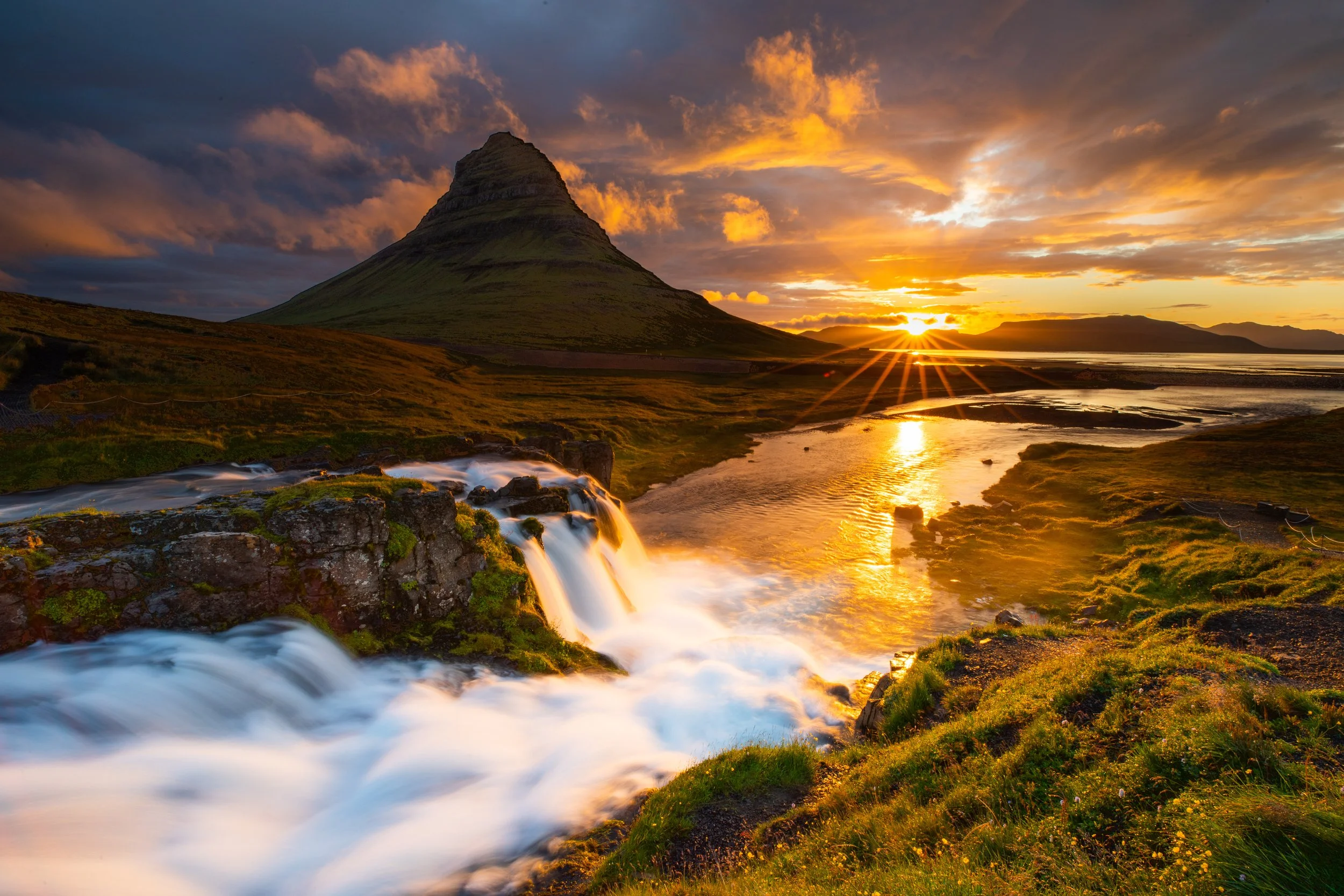 A scenic landscape of Kirkjufell mountain at sunset in Iceland with a waterfall in the foreground and a river reflecting the golden sky.