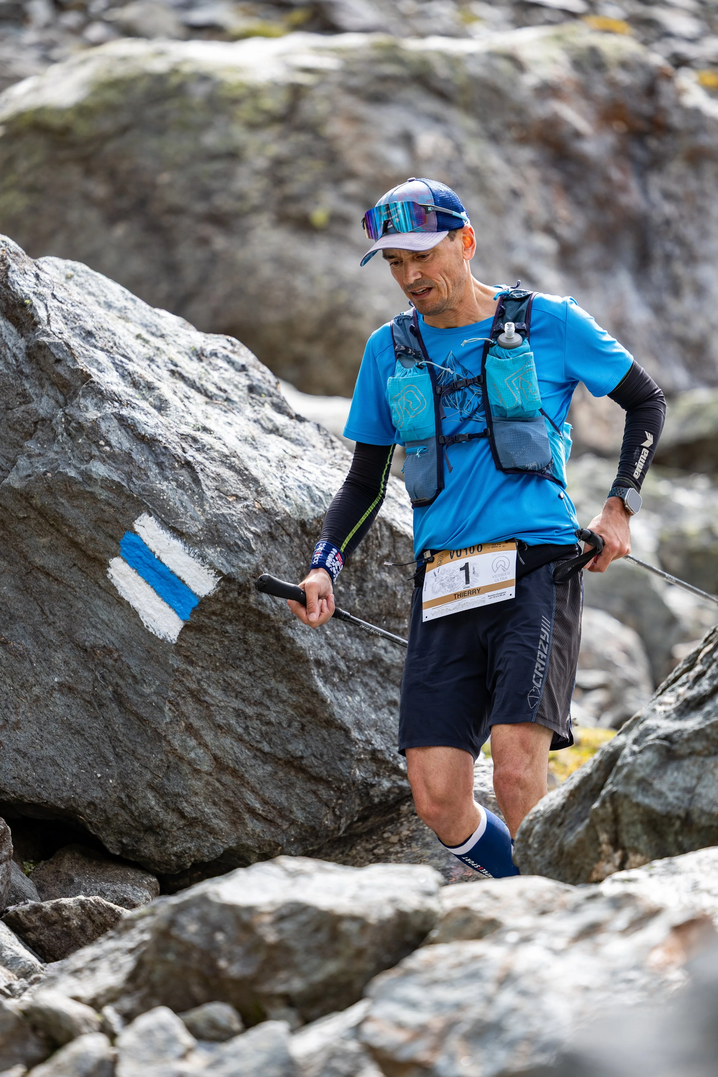 A man wearing a blue athletic shirt, black shorts, and a cap trails over rocky terrain with trekking poles, practicing trail running or mountain hiking.