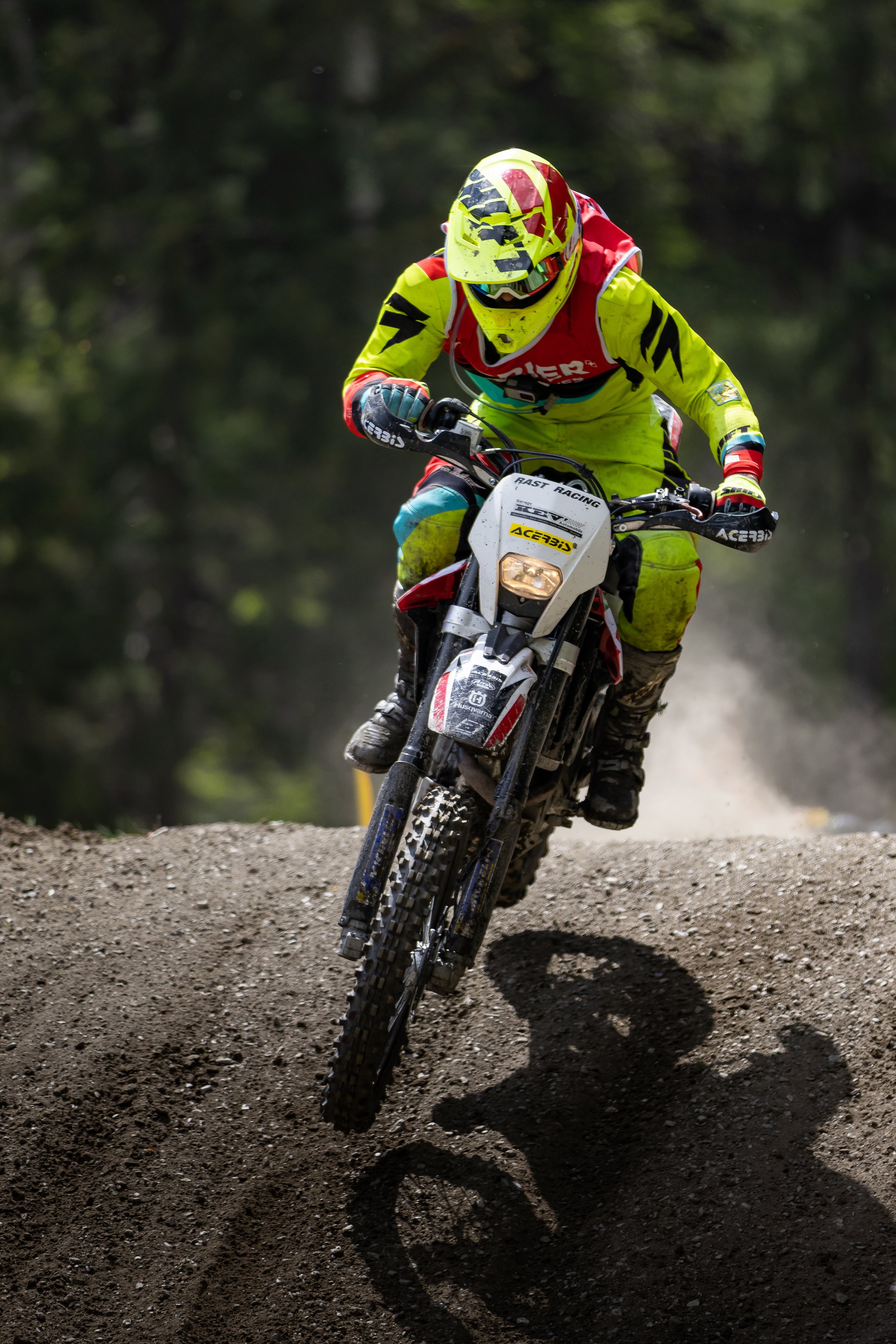 A motocross rider wearing a yellow and red helmet and neon yellow gear riding a dirt bike on a muddy trail surrounded by trees.