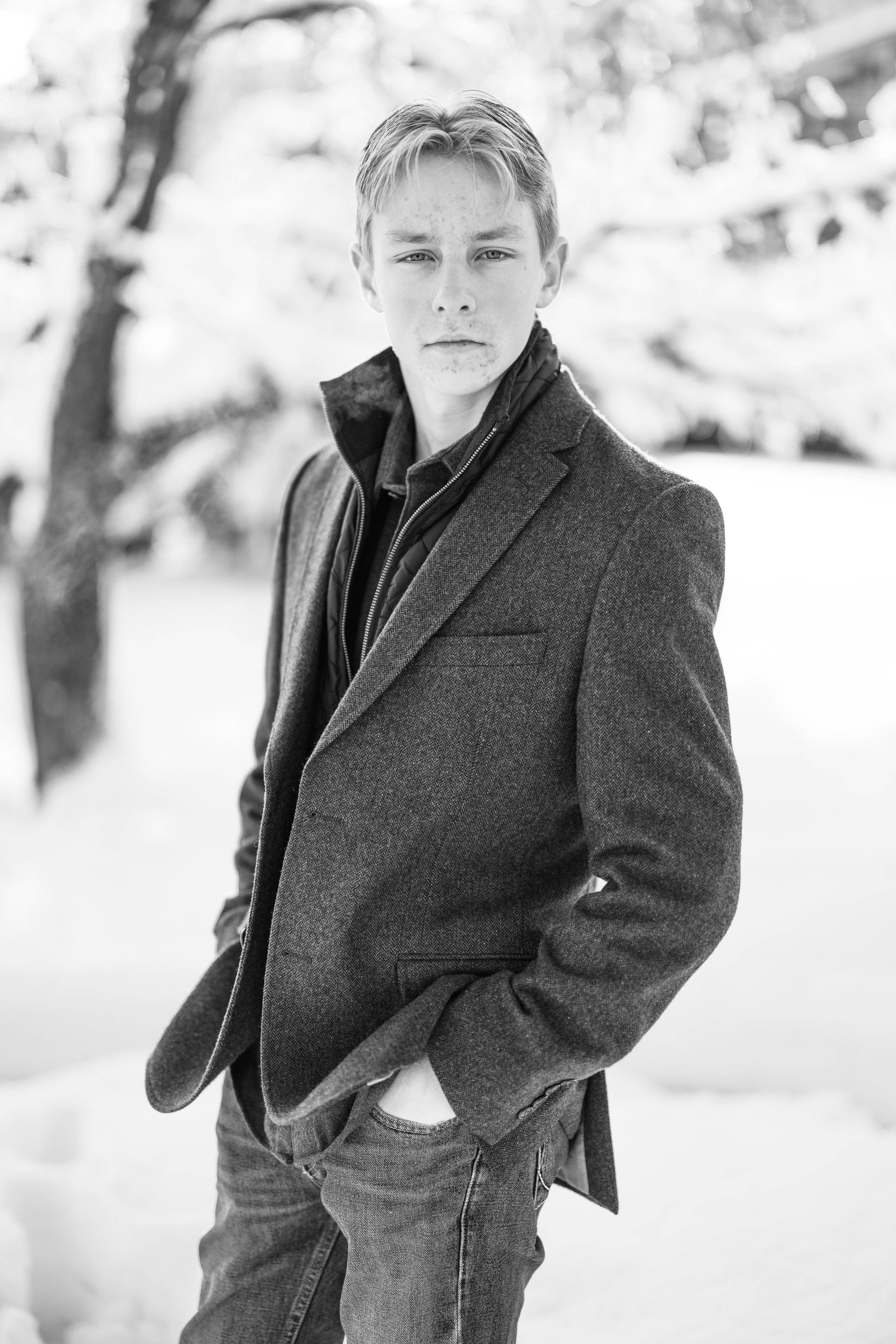 A young man standing outdoors in a snowy landscape, wearing a dark blazer and casual jeans, with a serious expression on his face.
