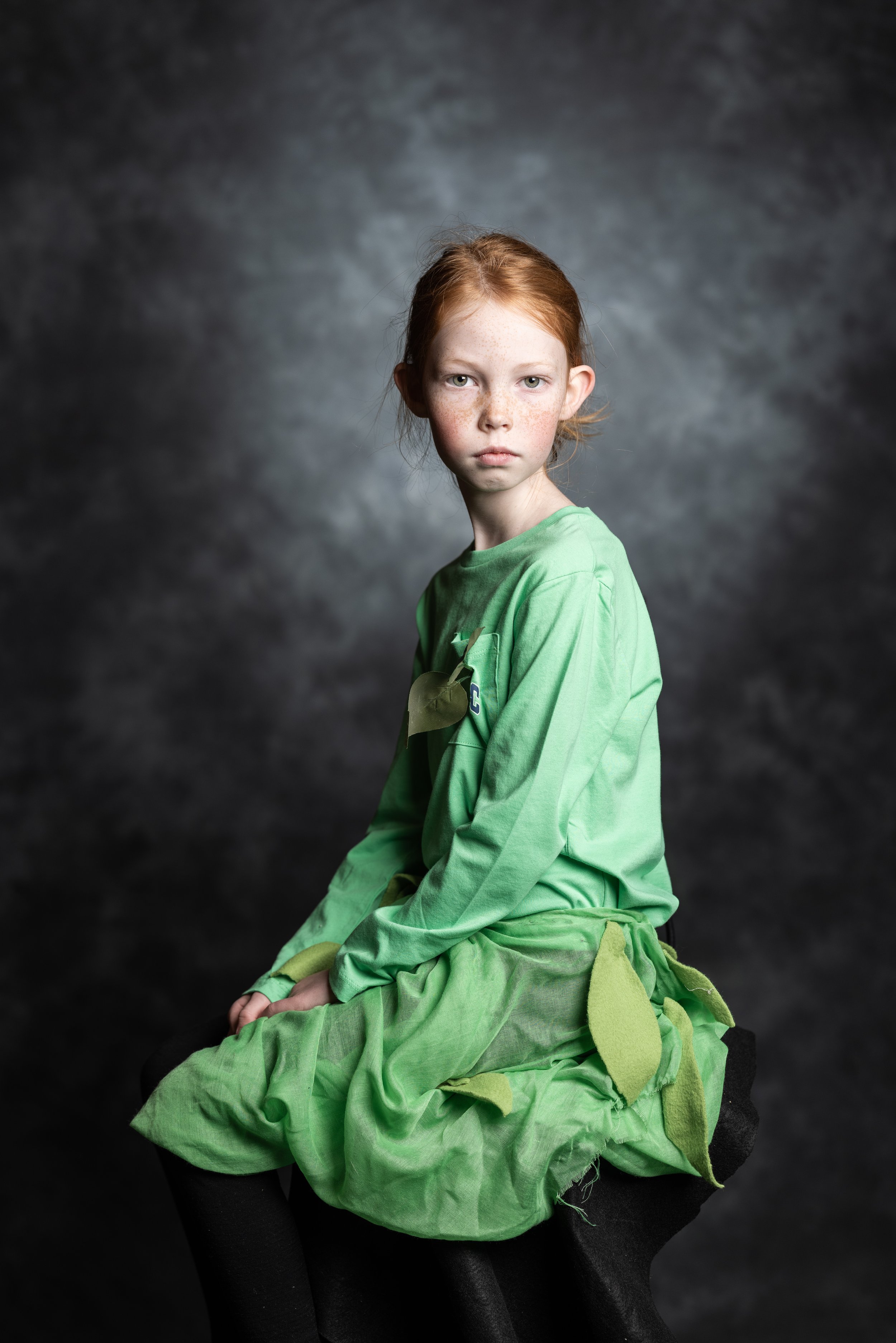 A young girl with red hair and freckles is sitting on a black stool against a dark gray textured background. She is wearing a bright green dress with leaf details.