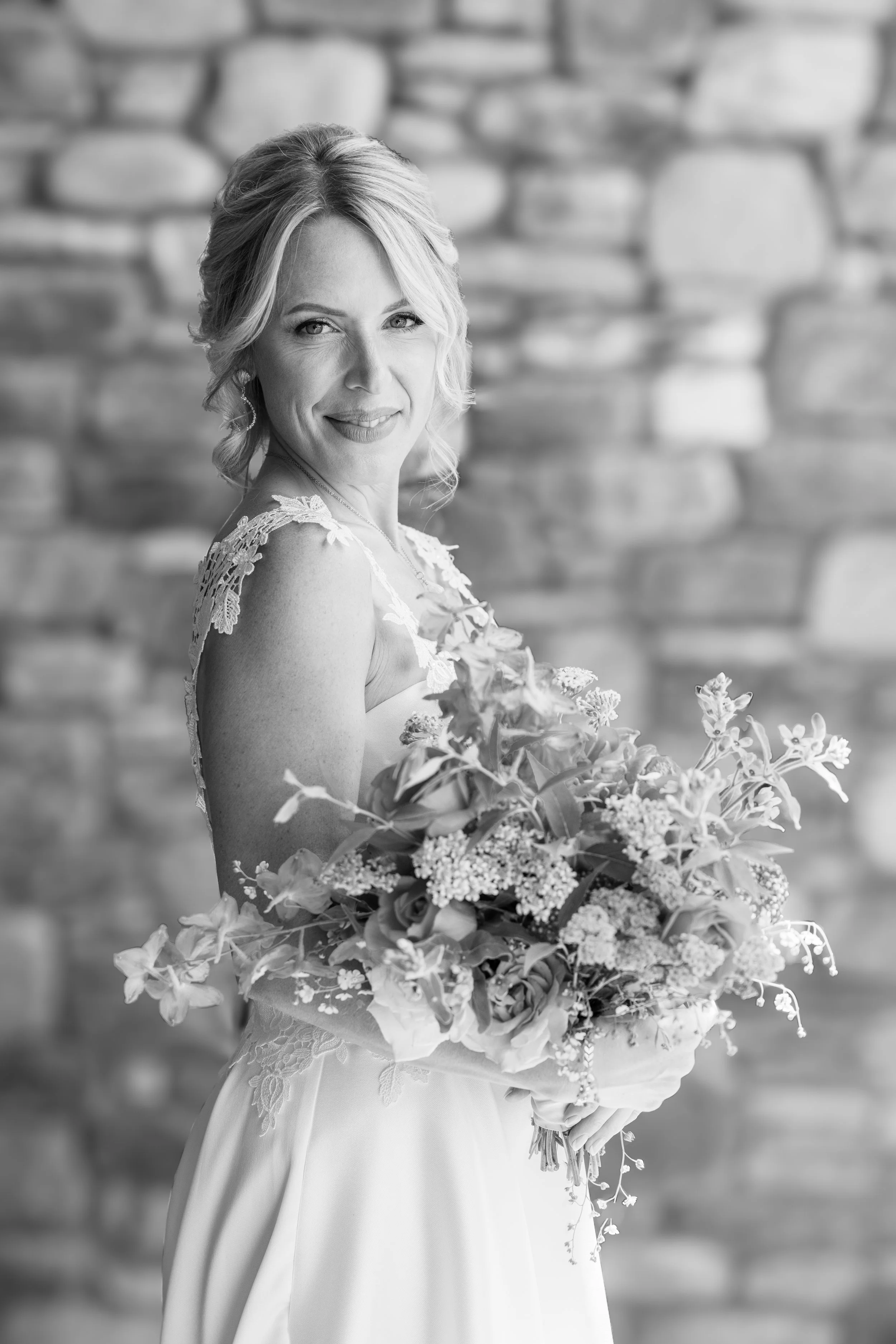 Black and white photo of a smiling woman in a wedding dress holding a bouquet of flowers in front of a stone wall taken at the Hôtel Cordée des Alpes, Verbier, Switzerland.