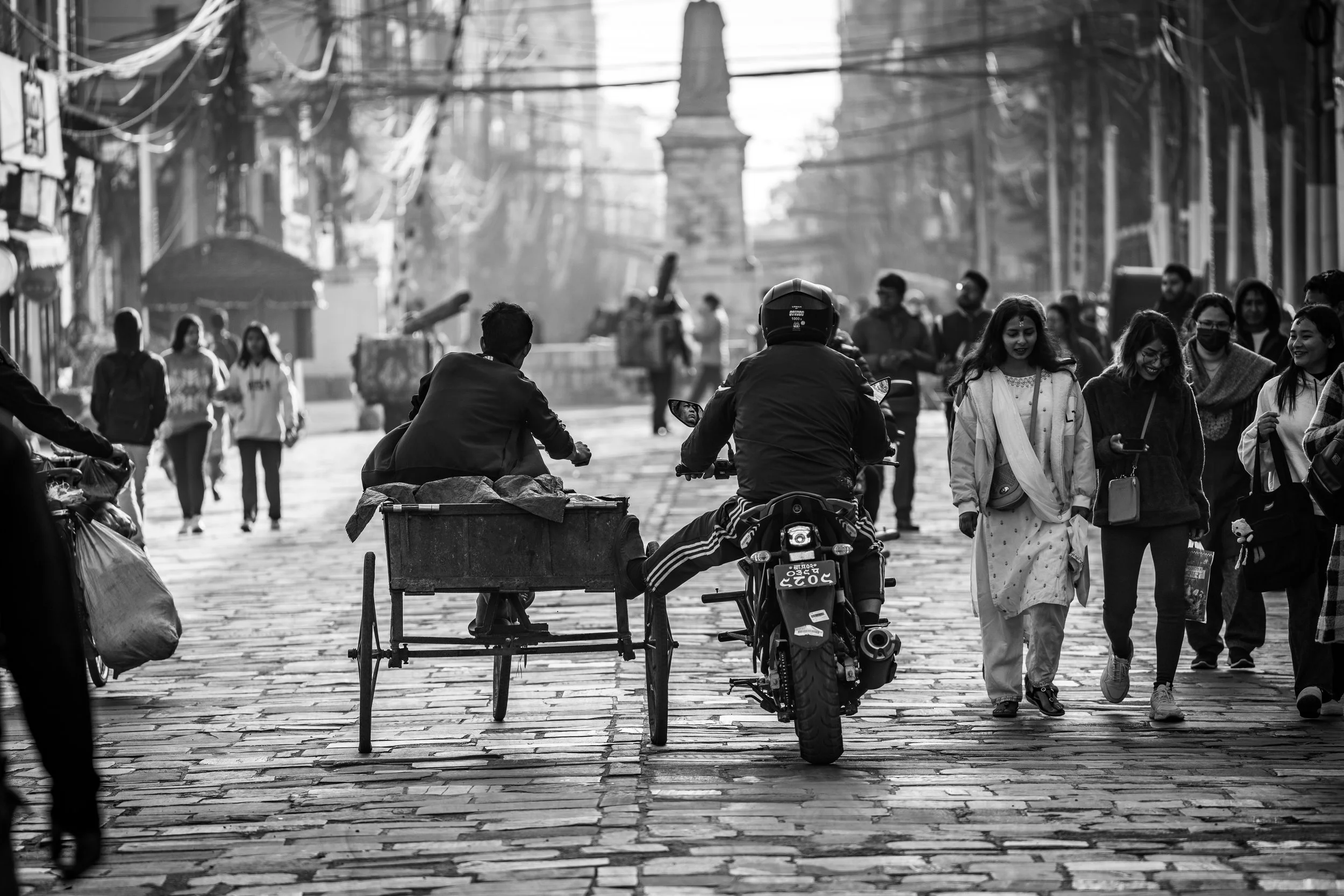 Black and white photo of a busy street scene with people walking and riding a motorcycle with a carrier sidecar. The street is paved with bricks, and there are buildings and electric wires overhead. A monument or statue is visible in the background.