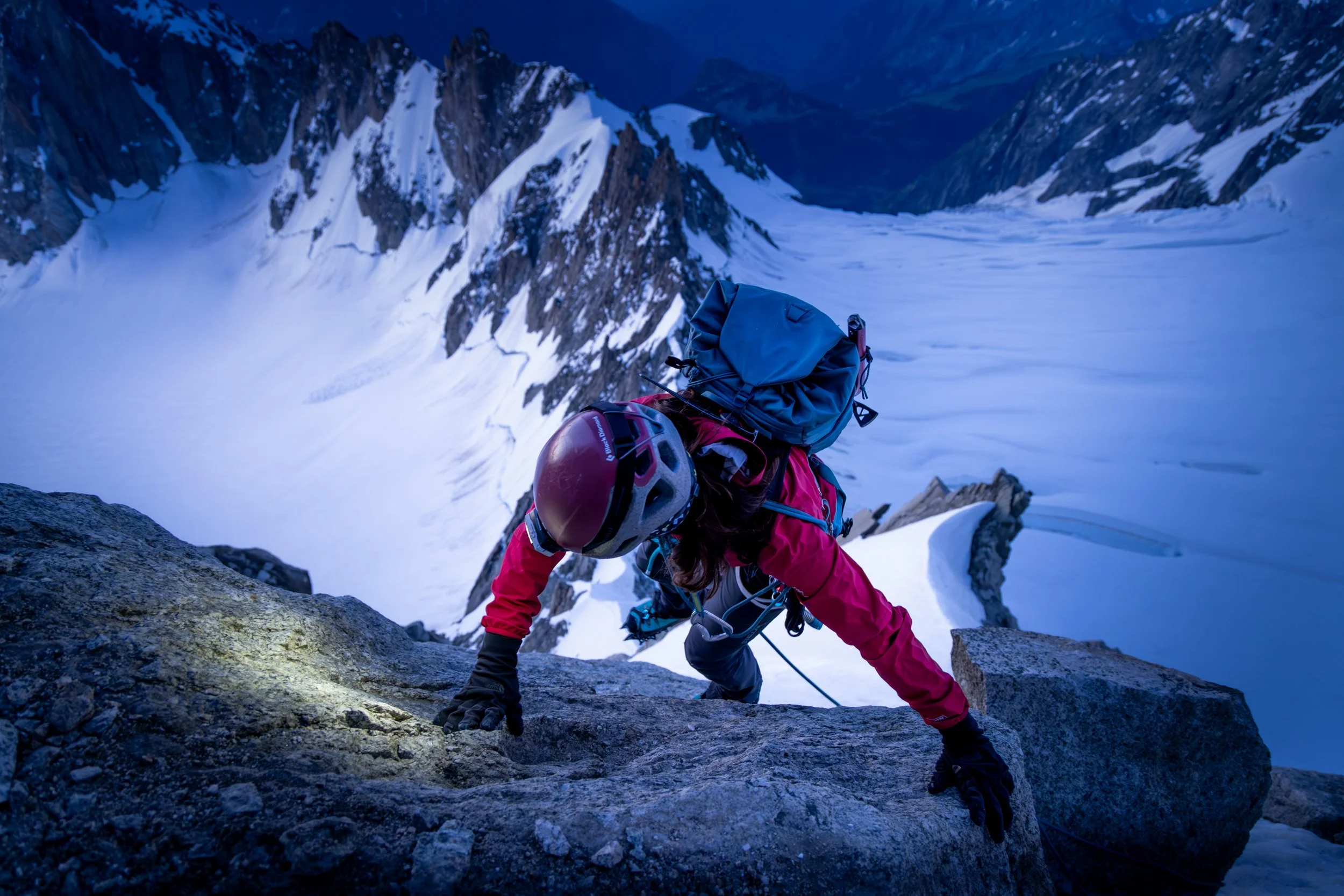 Climber in red jacket and helmet ascending snow-covered mountain ridge with icy landscape in background.