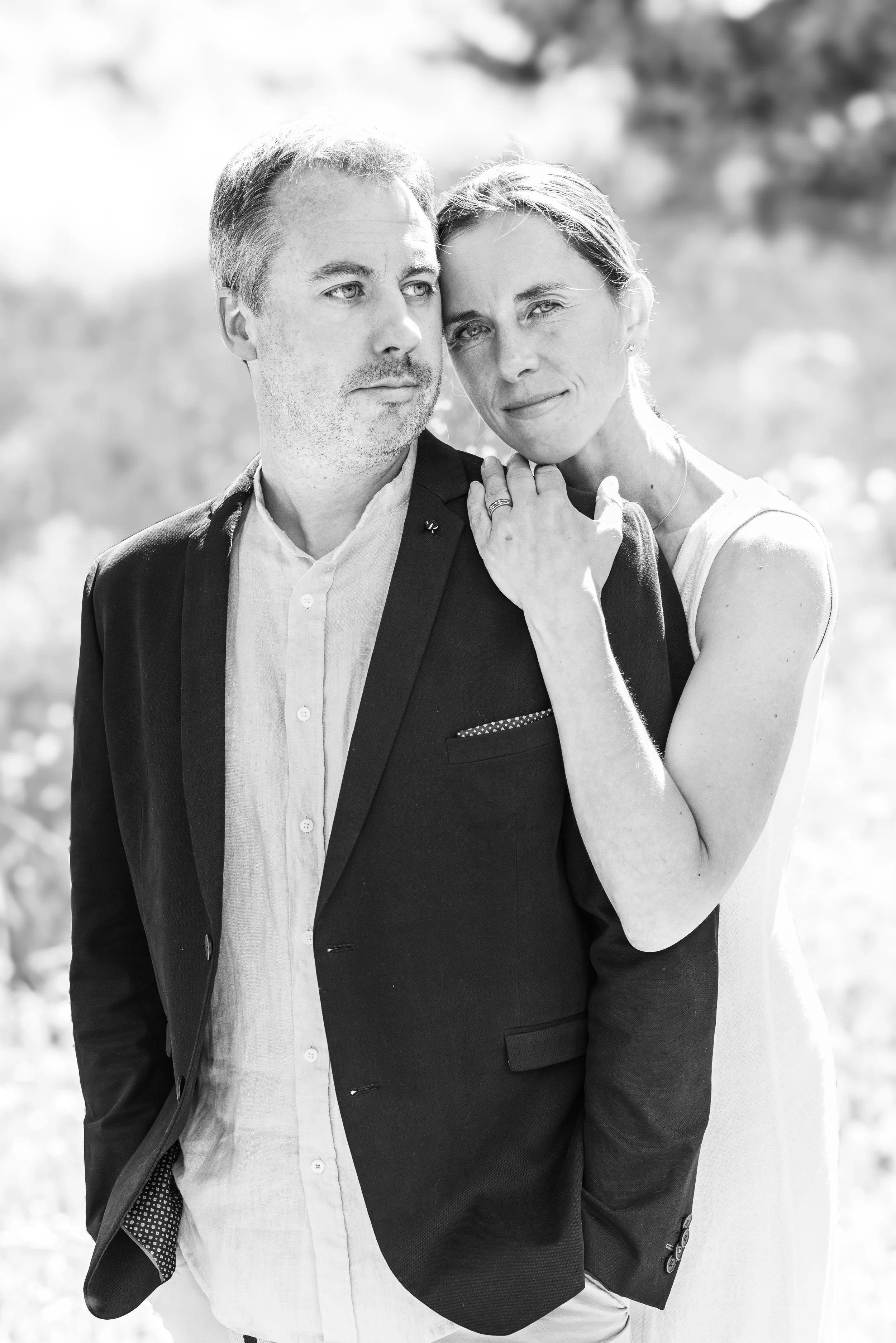 A black and white photograph of a couple outdoors, with the woman resting her head on the man's shoulder, both looking at the camera. The woman has her arm around the man's shoulder, with her hand on his neck, and the man is wearing a blazer over a s