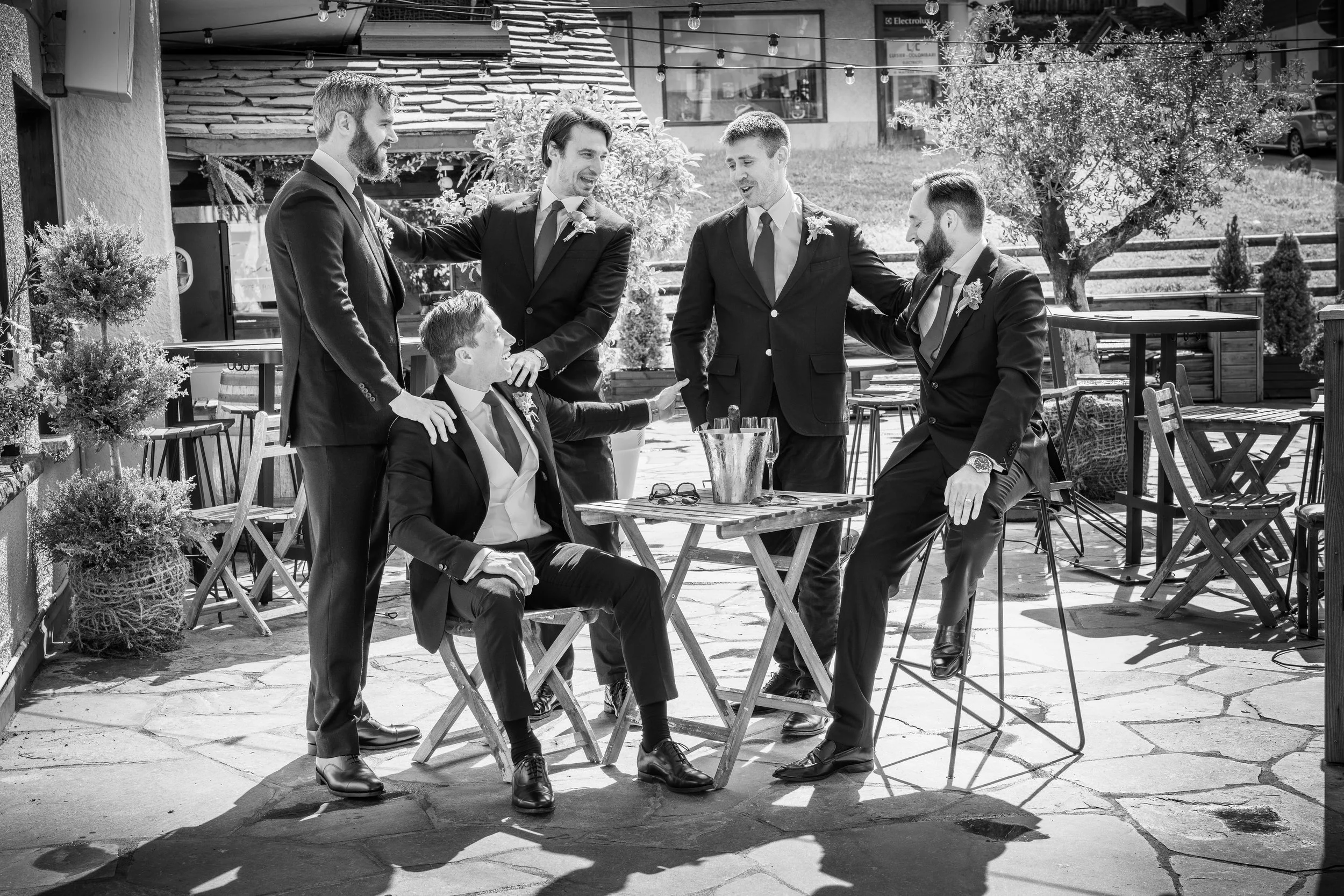 Six men in suits gathered around a table outdoors, laughing and talking, with some standing and some seated, in a casual social setting taken in The Farinet Hotel, Verbier, Switzerland.