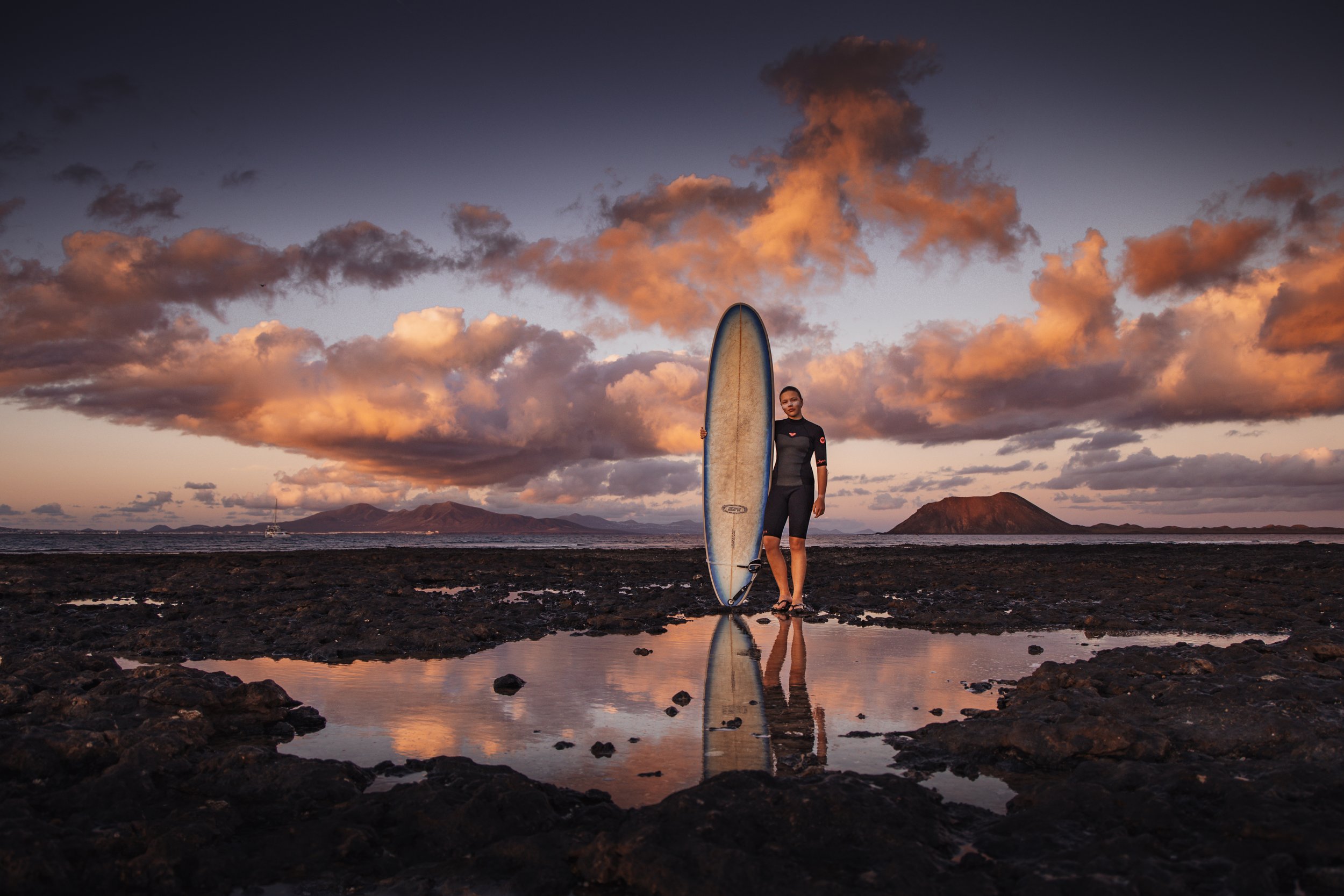 A woman standing on a rocky beach holding a surfboard during sunset, with clouds and mountains in the background.