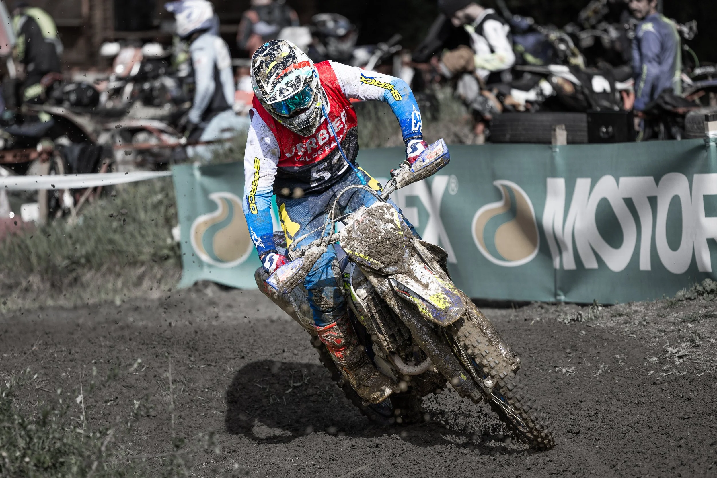 A motocross rider wearing a protective helmet and colorful racing gear riding through muddy track, with spectators watching behind a barrier.