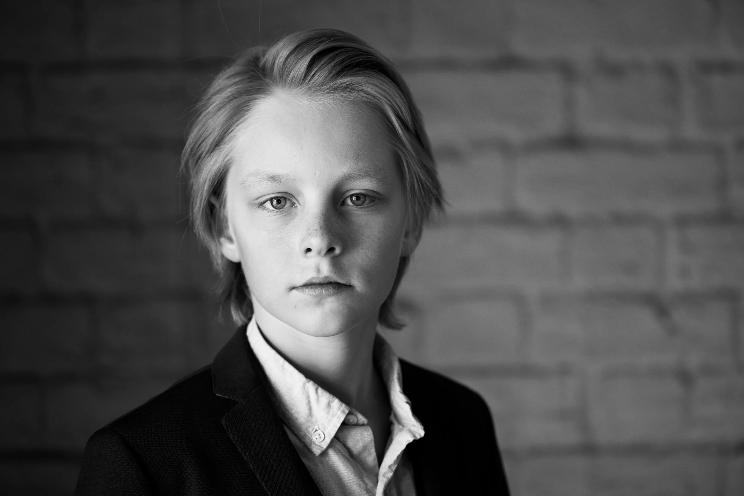 A young boy with shoulder-length blonde hair and light skin looking directly at the camera. He is wearing a dark blazer over a light shirt and standing in front of a brick wall.