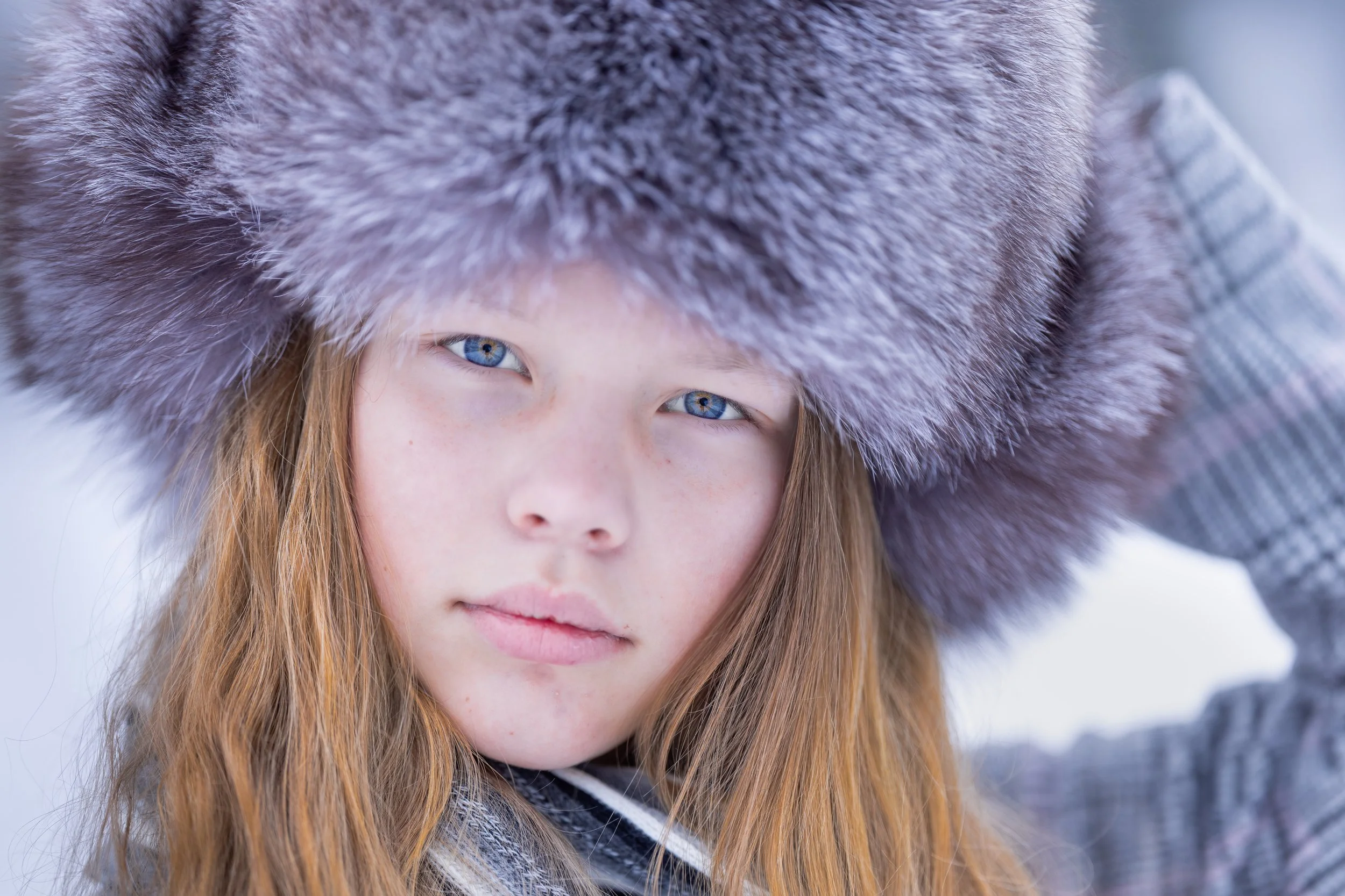 Close-up of a young girl with blue eyes and long red hair wearing a large fuzzy lavender hat and a plaid jacket, outdoors in a snowy setting.