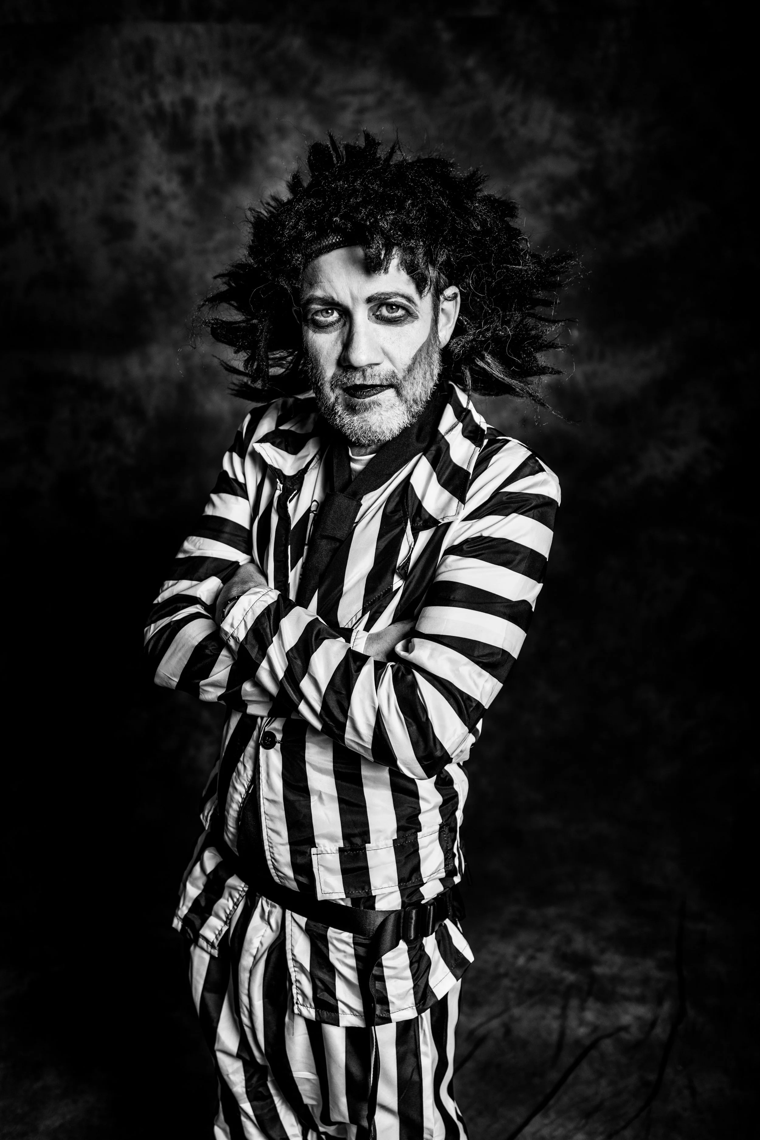Black and white portrait of a man dressed as a mime with striped clothing and wild hair, posing with arms crossed, against a dark background.
