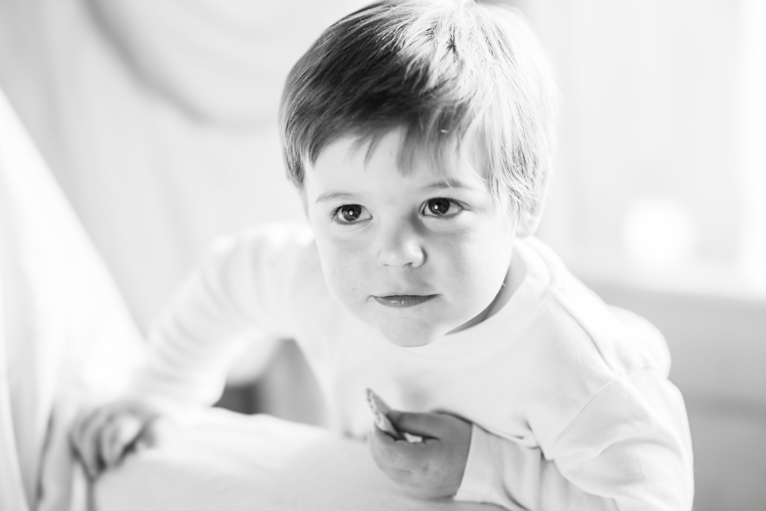 A young boy with short hair looking up at the camera. He is leaning over a surface, wearing a white shirt, and holding something in his hand.