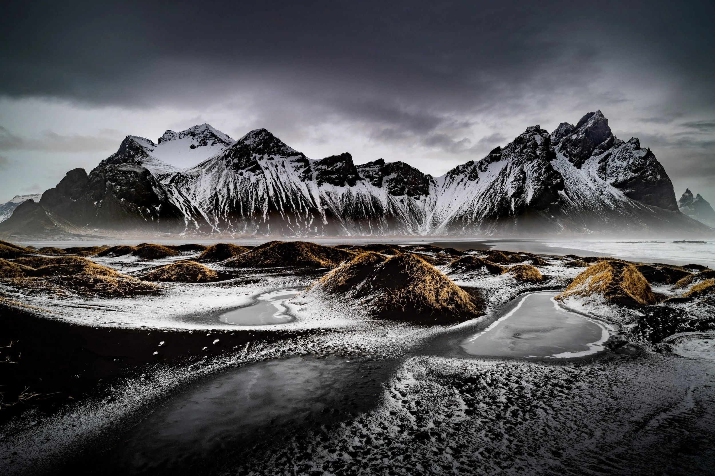 Snow-capped mountains in the background with dark, cloudy sky, and black volcanic sand beach in the foreground with small pools of water and patches of dried grass.
