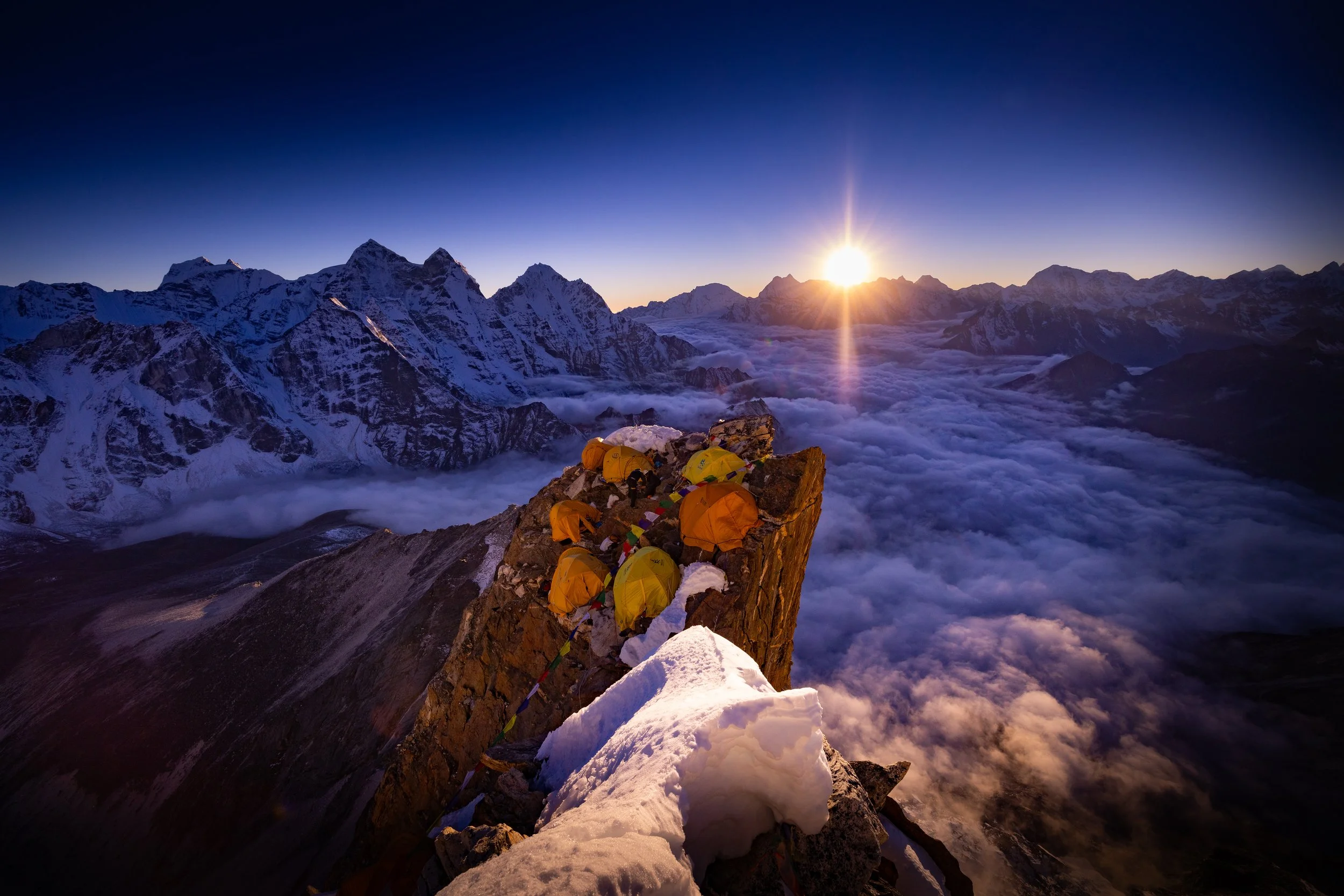 A group of climbers with yellow tents on a snow-covered mountain ridge during sunrise, with snow-capped peaks and a sea of clouds below.