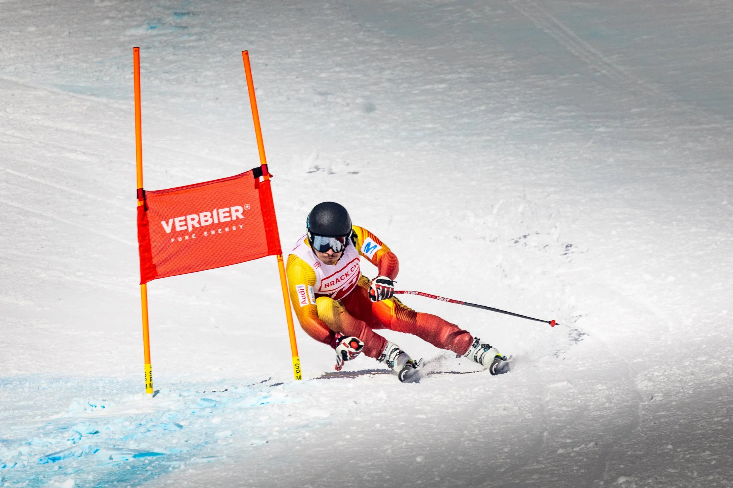 A skier in a racing suit, helmet, and goggles is leaning into a turn on snow, passing a red gate with 'VERBIER PURE ENERGY' written on it.