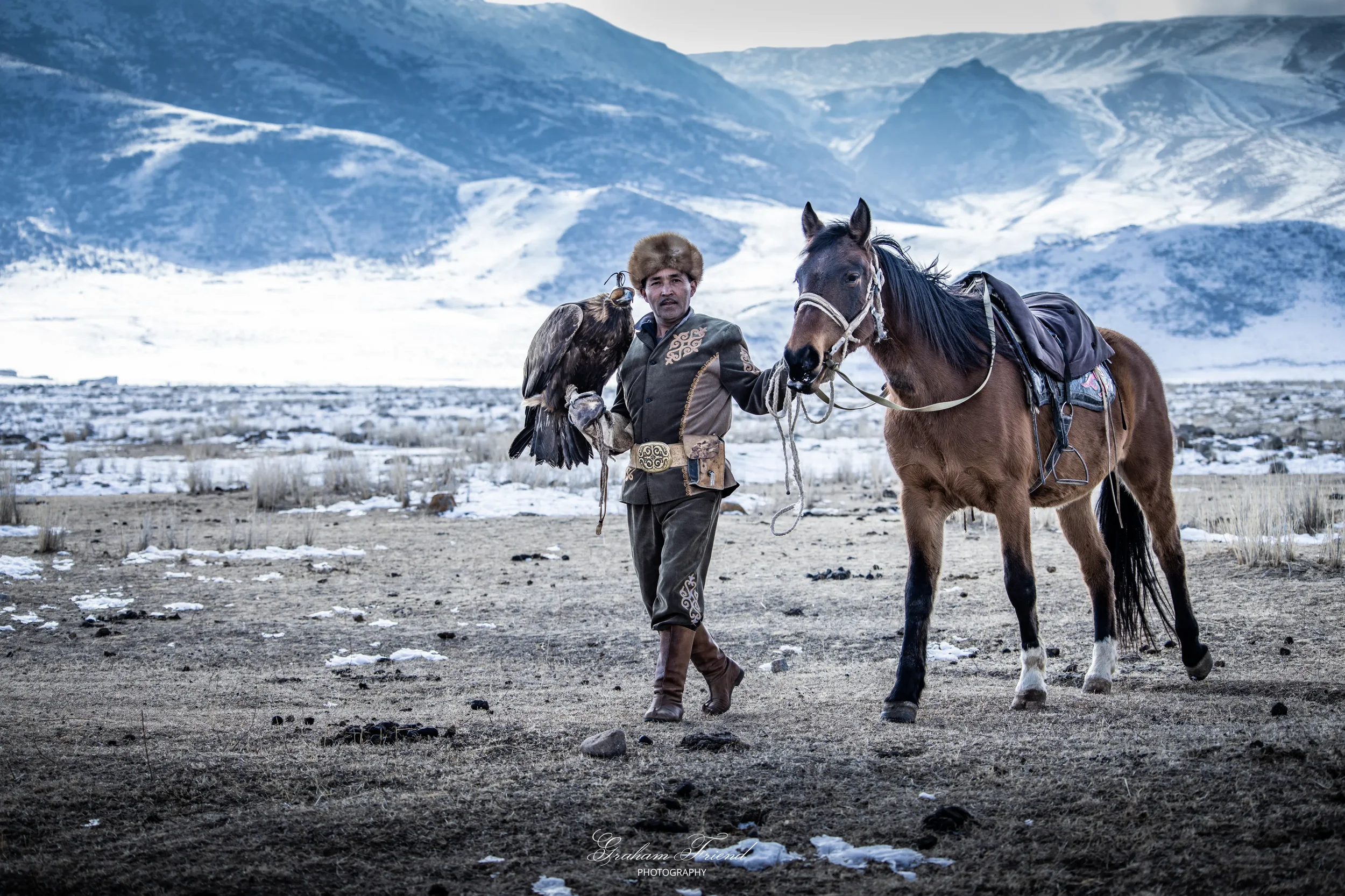 A man wearing traditional Mongolian clothing holding a bird of prey on his arm, standing next to a brown horse in a snowy mountain landscape.