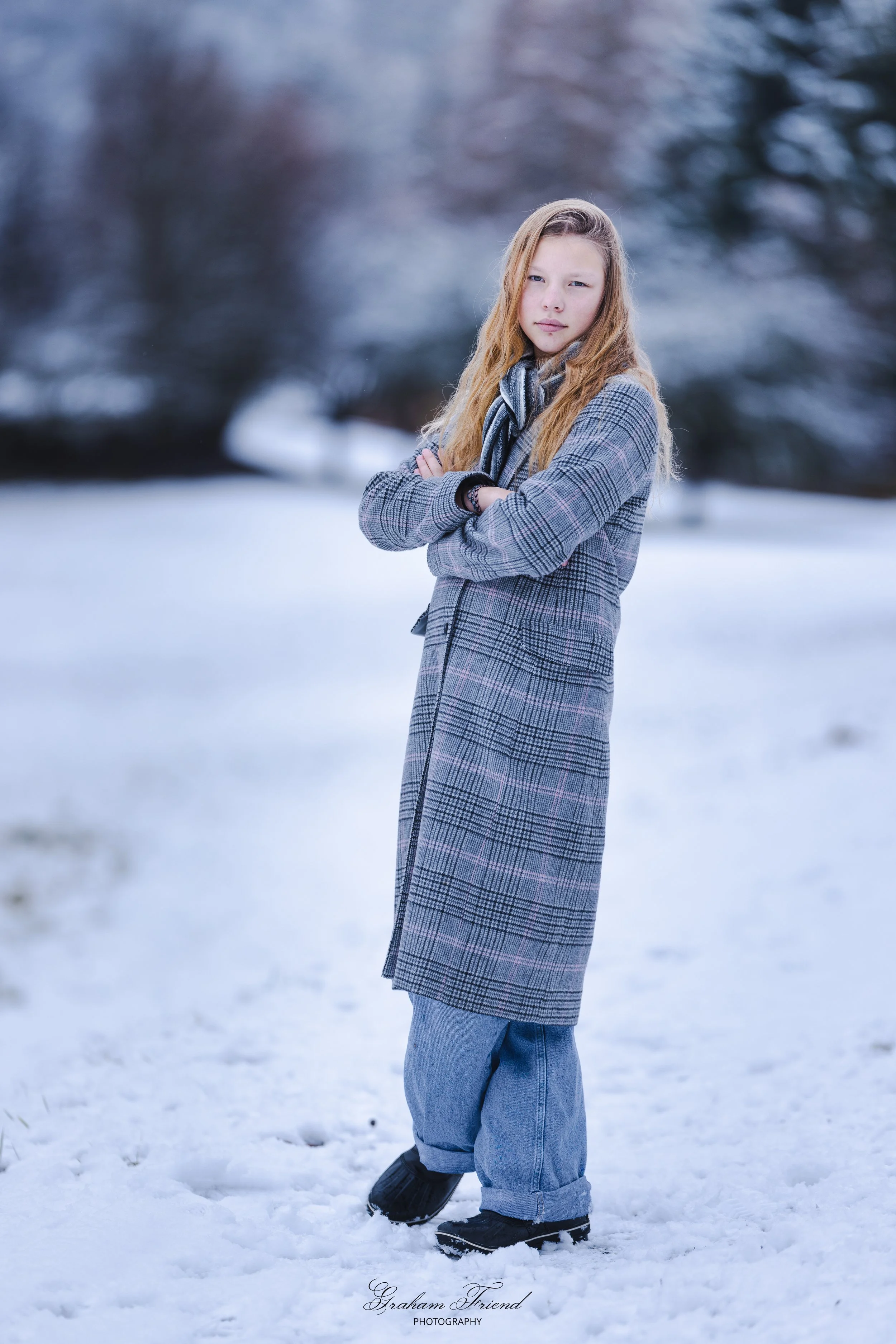 A young girl with long blonde hair, arms crossed, standing in a snowy outdoor setting with blurred trees in the background, wearing a gray plaid coat, blue jeans, and black boots.