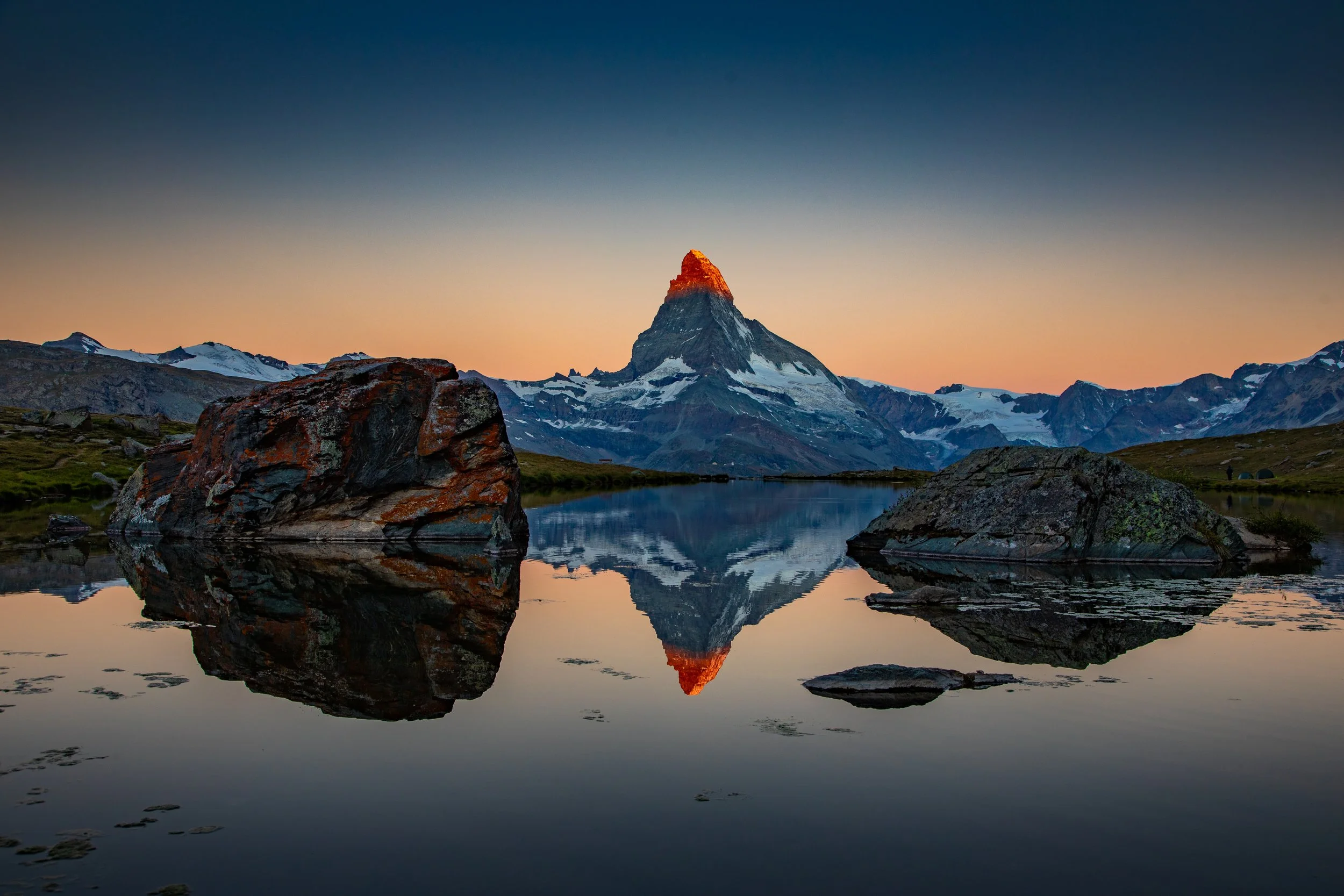 Mountaintop reflected in a calm lake with large rocks in the foreground, snow-capped peaks, and a colorful sky at sunrise or sunset.