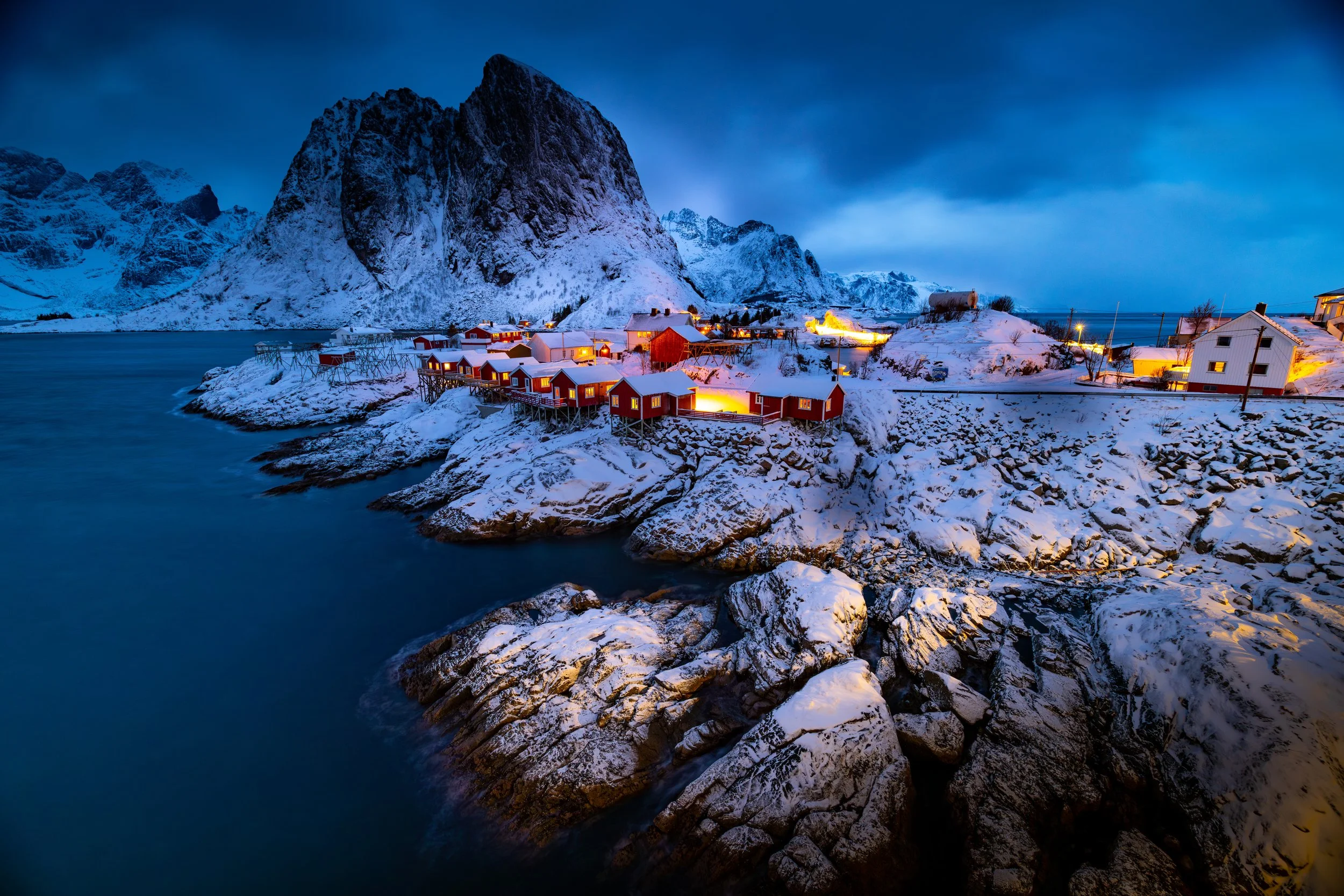 Snow-covered small houses and buildings illuminated with warm lights on rocky coastline, with majestic snow-capped mountains in background under evening sky.