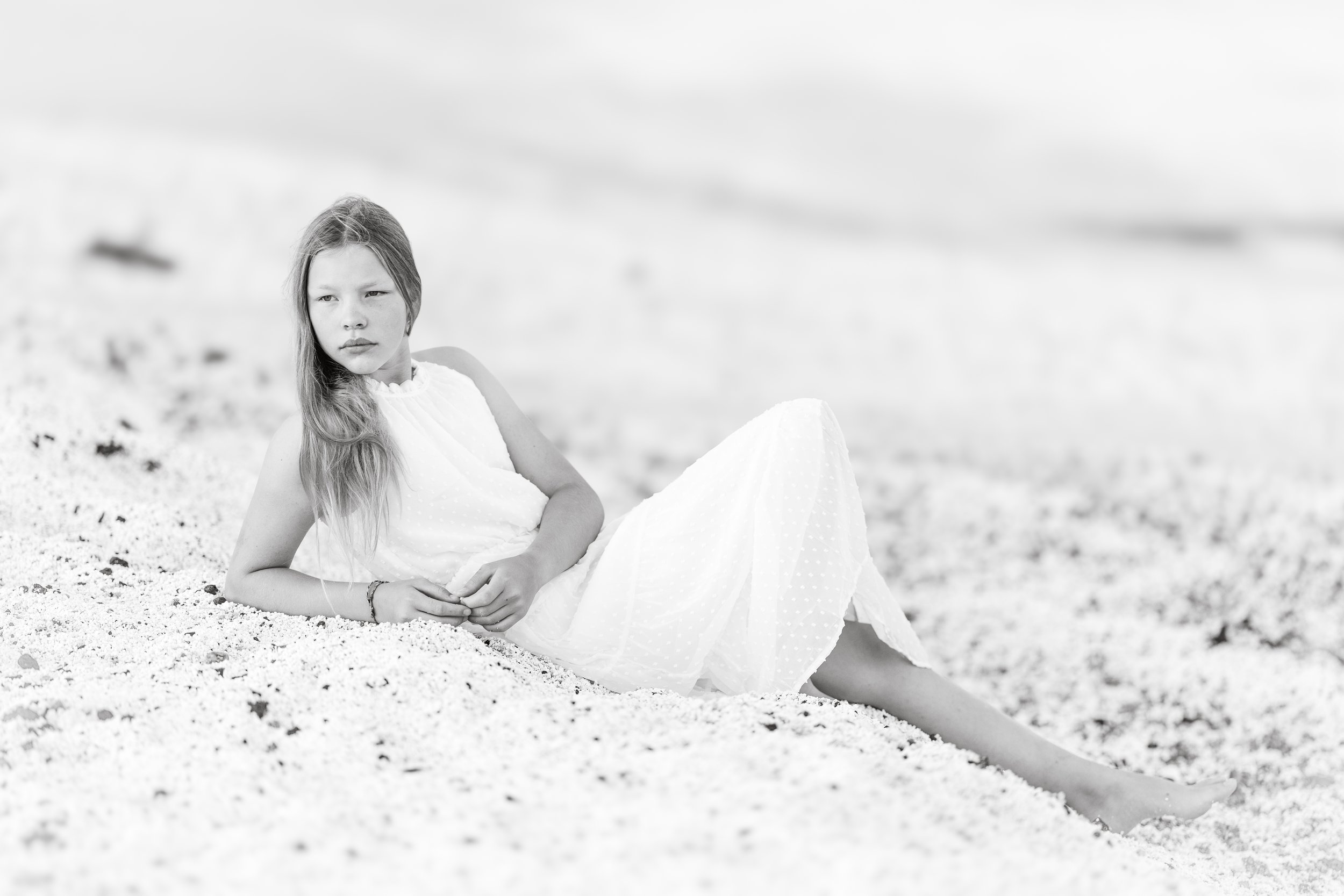 Black and white photo of a girl with long hair lying on a sandy beach in a white dress, looking pensively to the left.