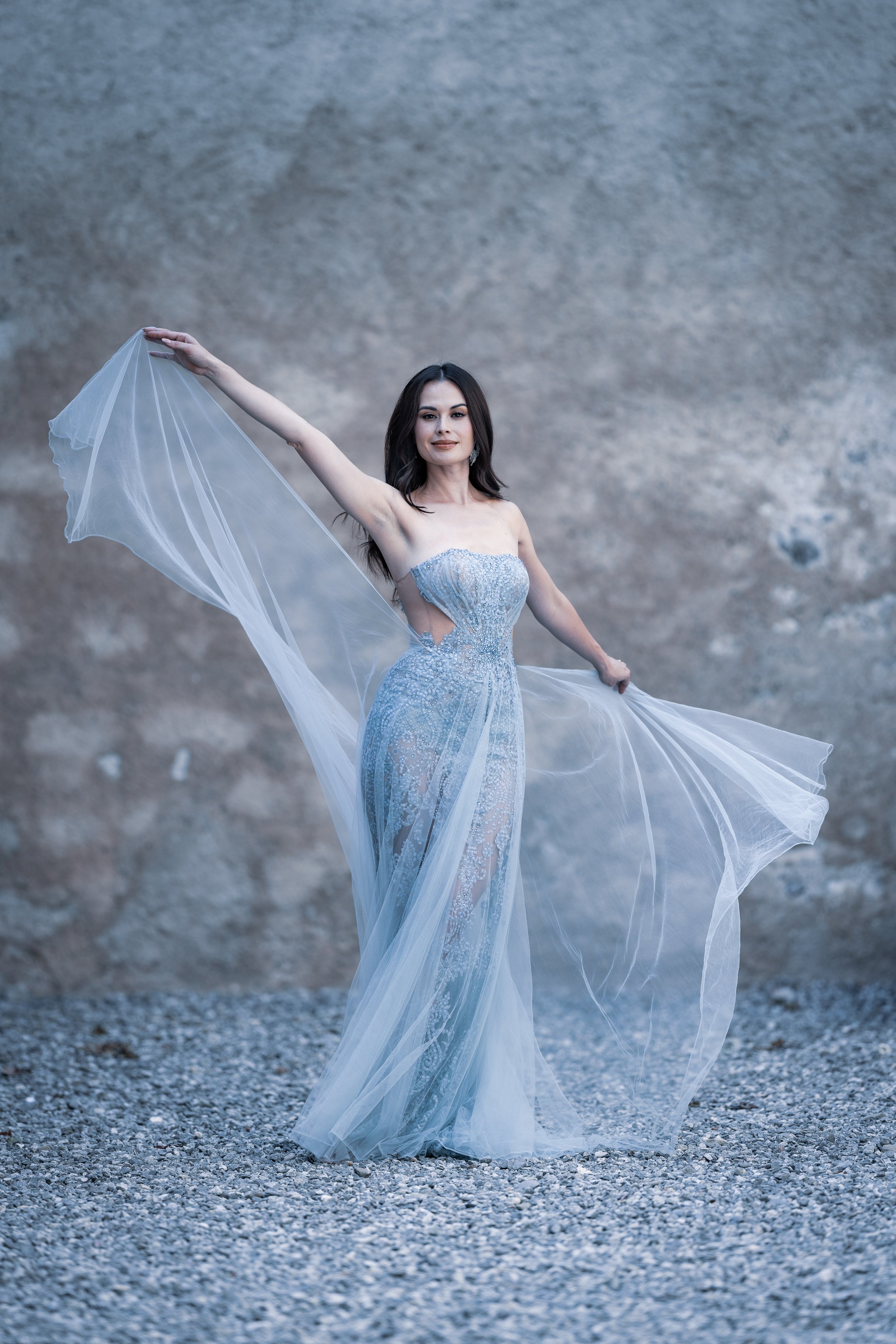 Woman in a sheer, blue, strapless gown with cutouts, dancing outdoors on gravel, arms extended, holding flowing fabric, against a textured wall background.