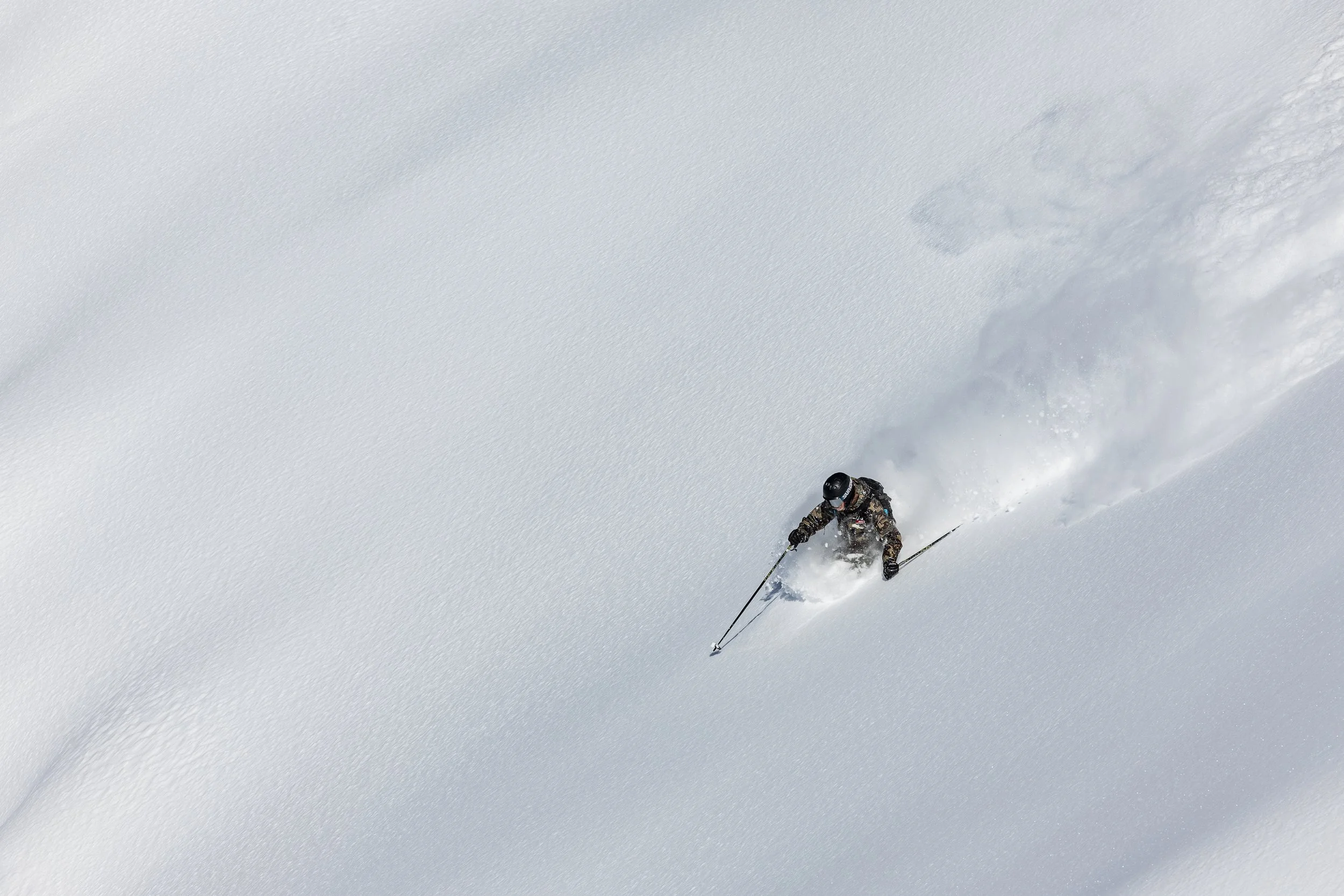 A skier in camouflage gear skiing down a snowy slope, creating a spray of snow.