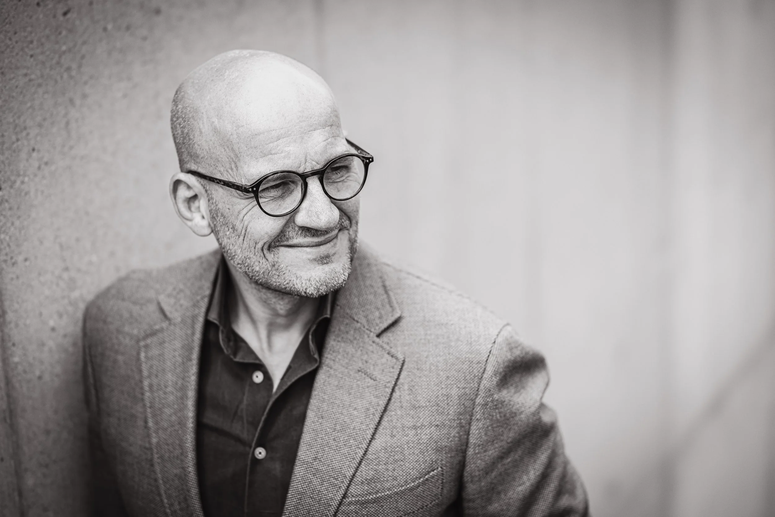 Black and white portrait of a bald man with glasses, smiling, wearing a blazer and shirt, leaning against a textured wall.