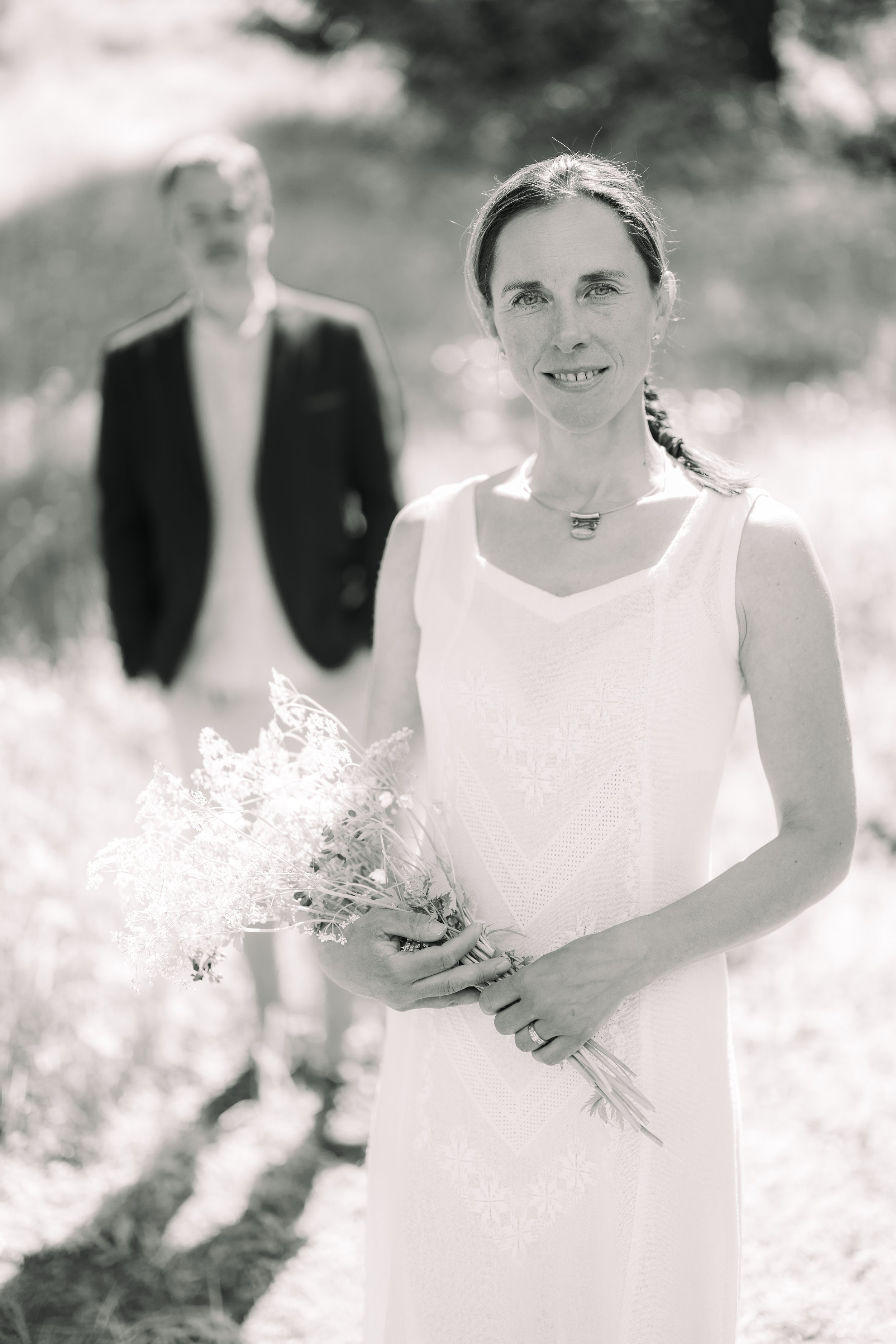 A woman in a white dress holding a bouquet of flowers outdoors, with a man in a dark suit blurred in the background.