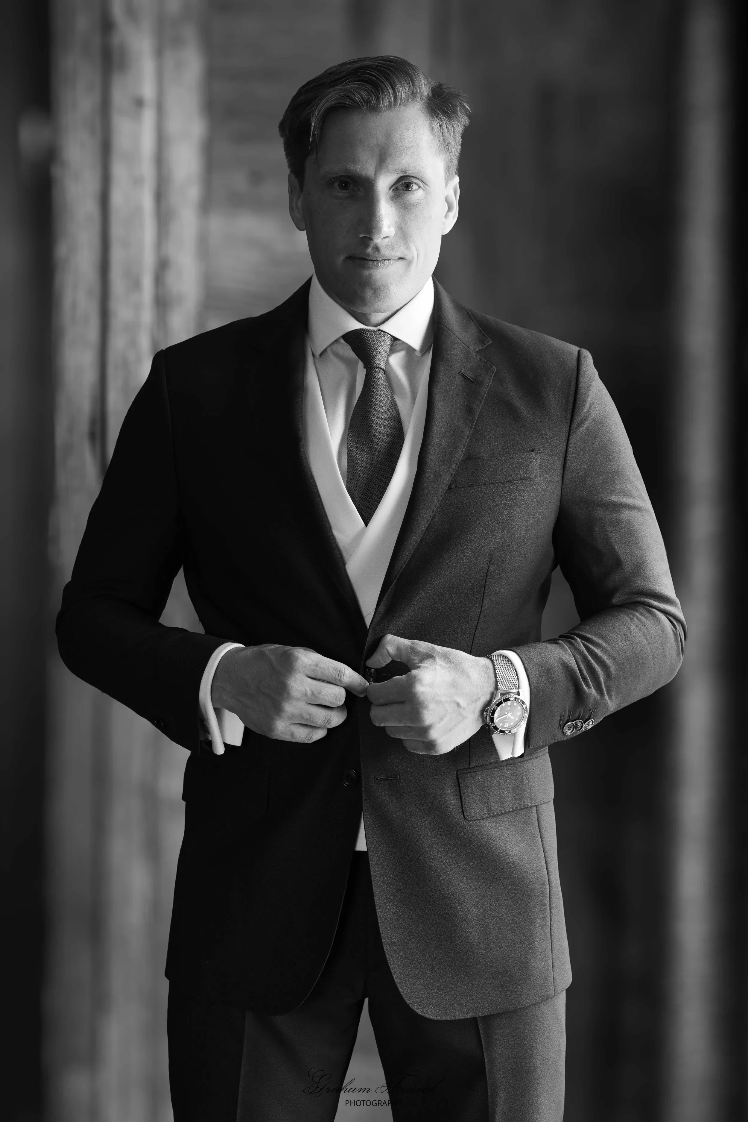 A black and white photo of a man in a suit, buttoning his jacket, standing indoors with a wooden wall background.