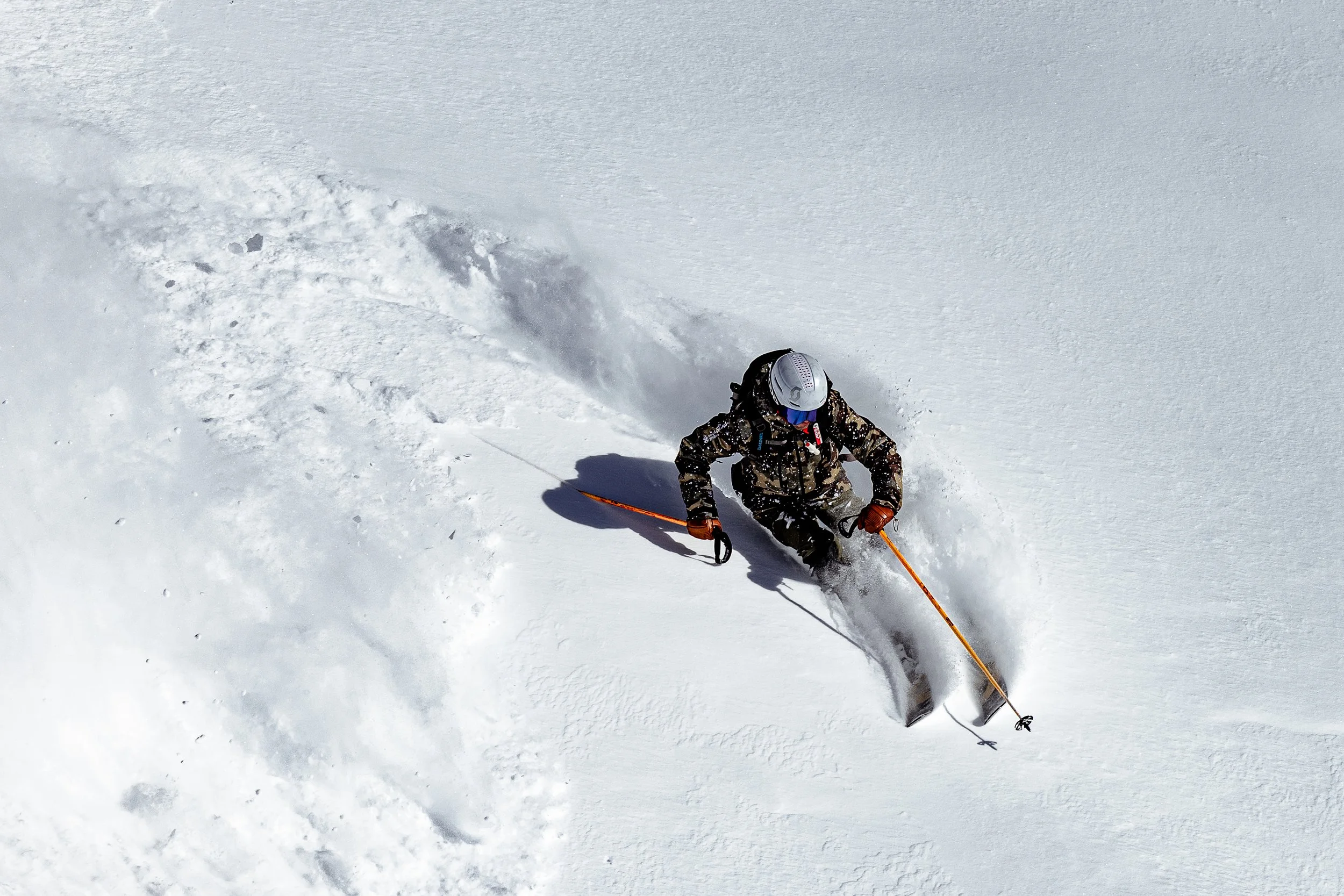 A person wearing winter gear, including a helmet and camouflage jacket, skiing down a snowy slope with tracks and snow spray.