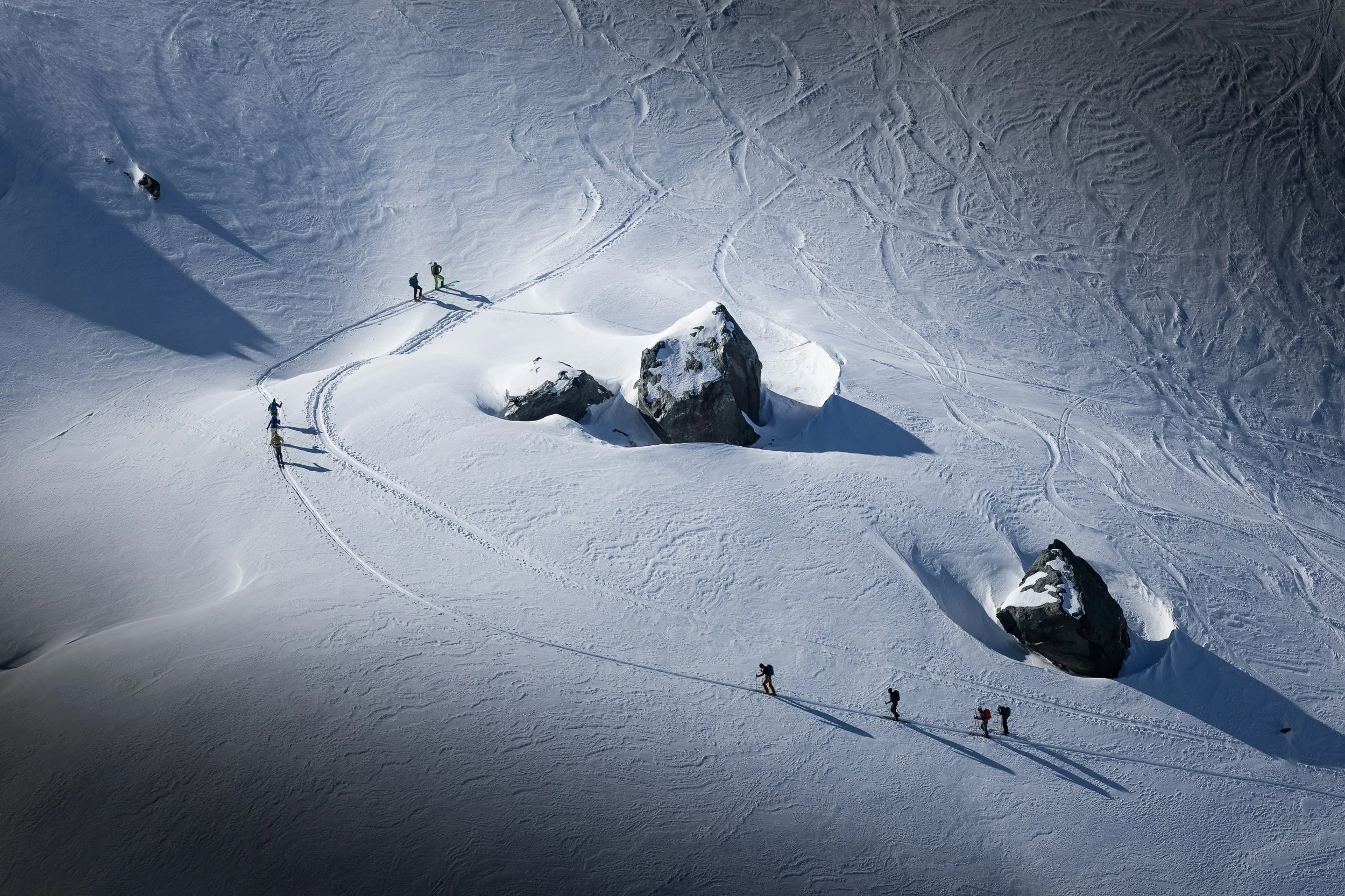 Aerial view of several skiers making their way down a snowy mountain around large rocks, with tracks and shadows visible on the snow.