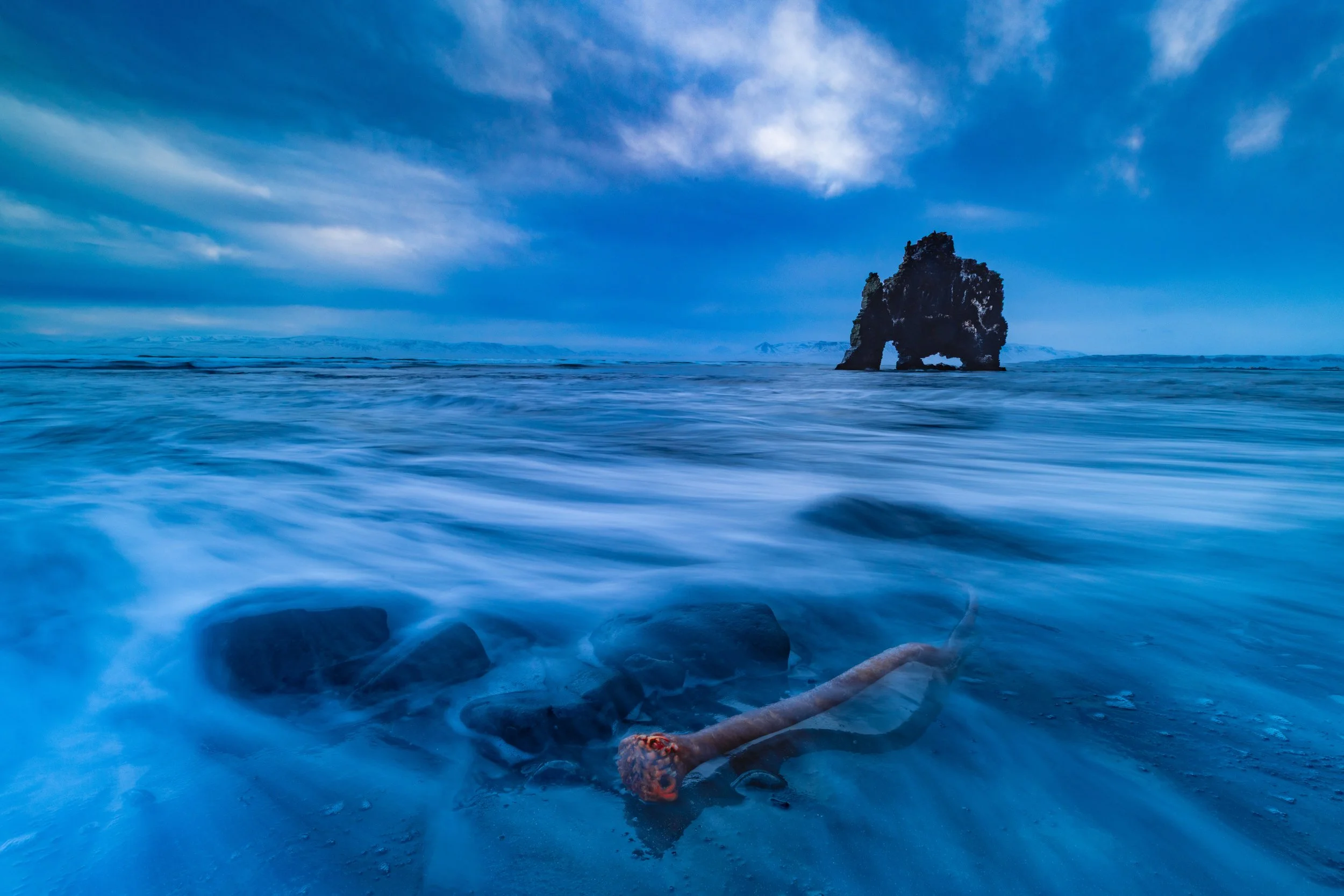 A rocky island formation in the ocean under cloudy sky, with a beached octopus on the shore in the foreground.