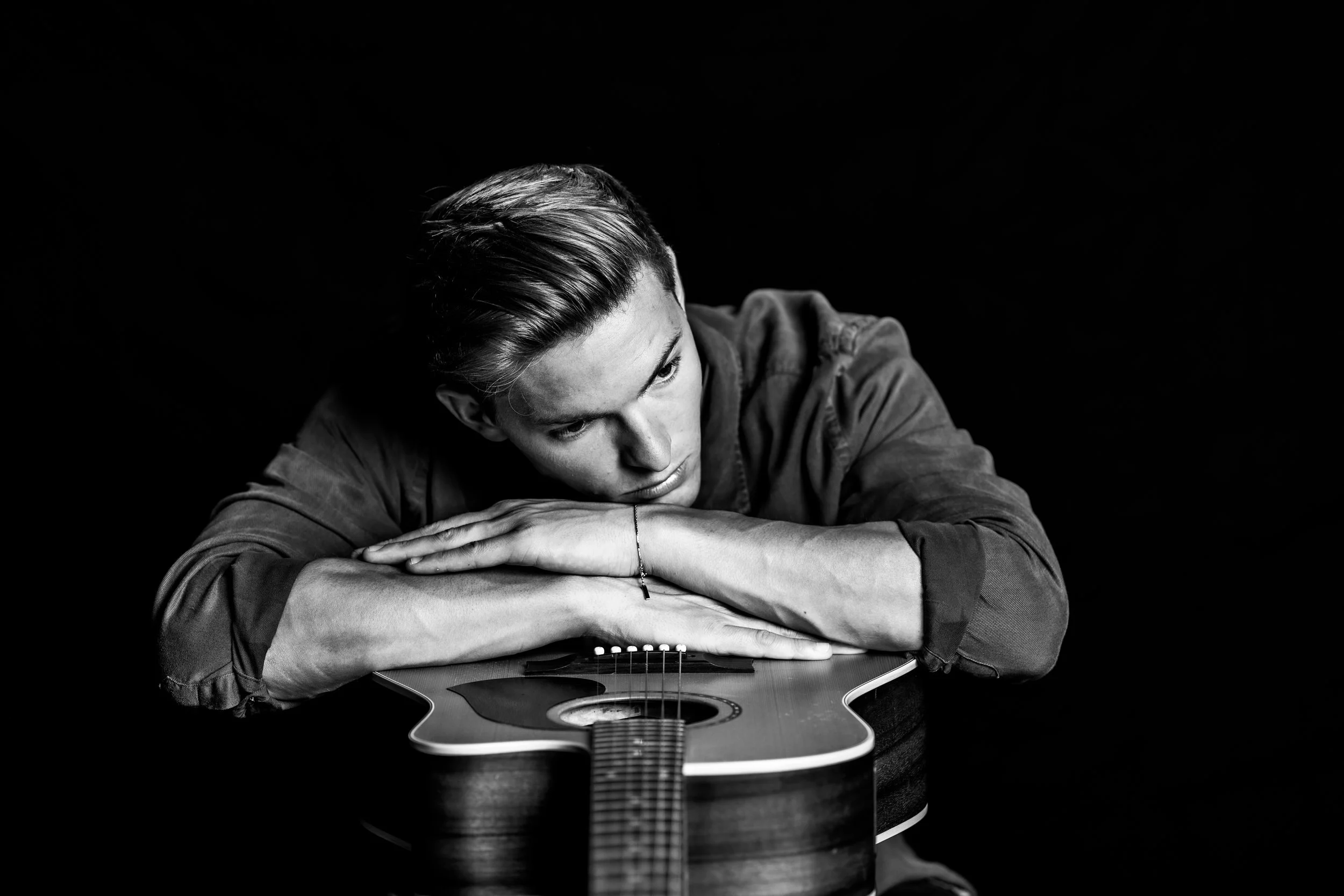 Black and white photo of a young man resting his head on an acoustic guitar, with the guitar lying flat on a dark surface. The man has short, slicked-back hair and is wearing a long-sleeved shirt.