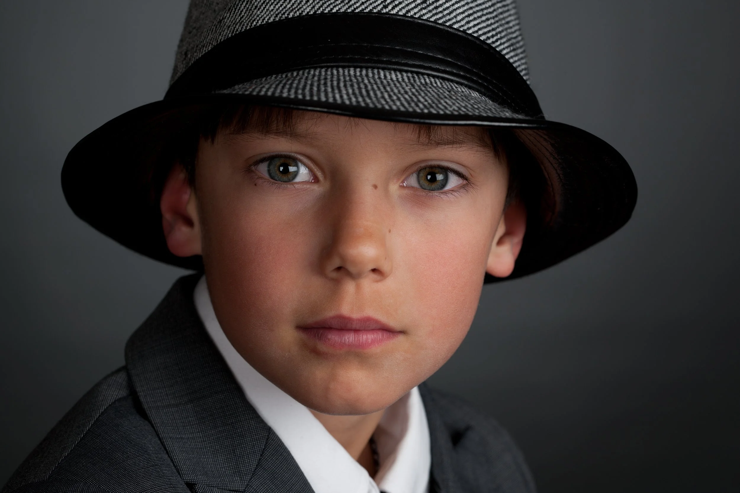 Close-up of a young boy with blue eyes wearing a gray suit, white shirt, and a black and gray fedora hat, against a dark background.
