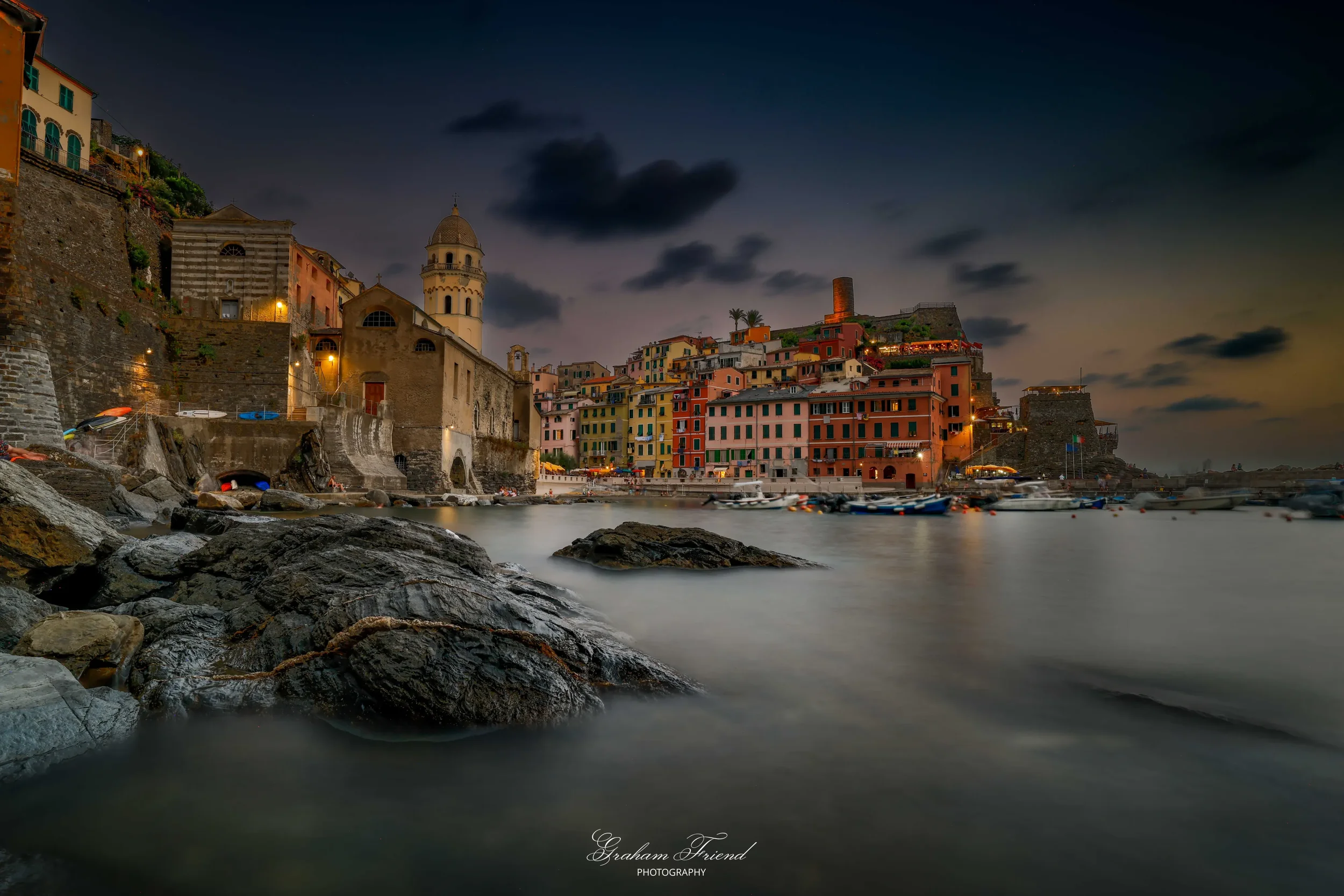 Colorful buildings along a harbor at dusk with boats anchored in the water and dark clouds in the sky.