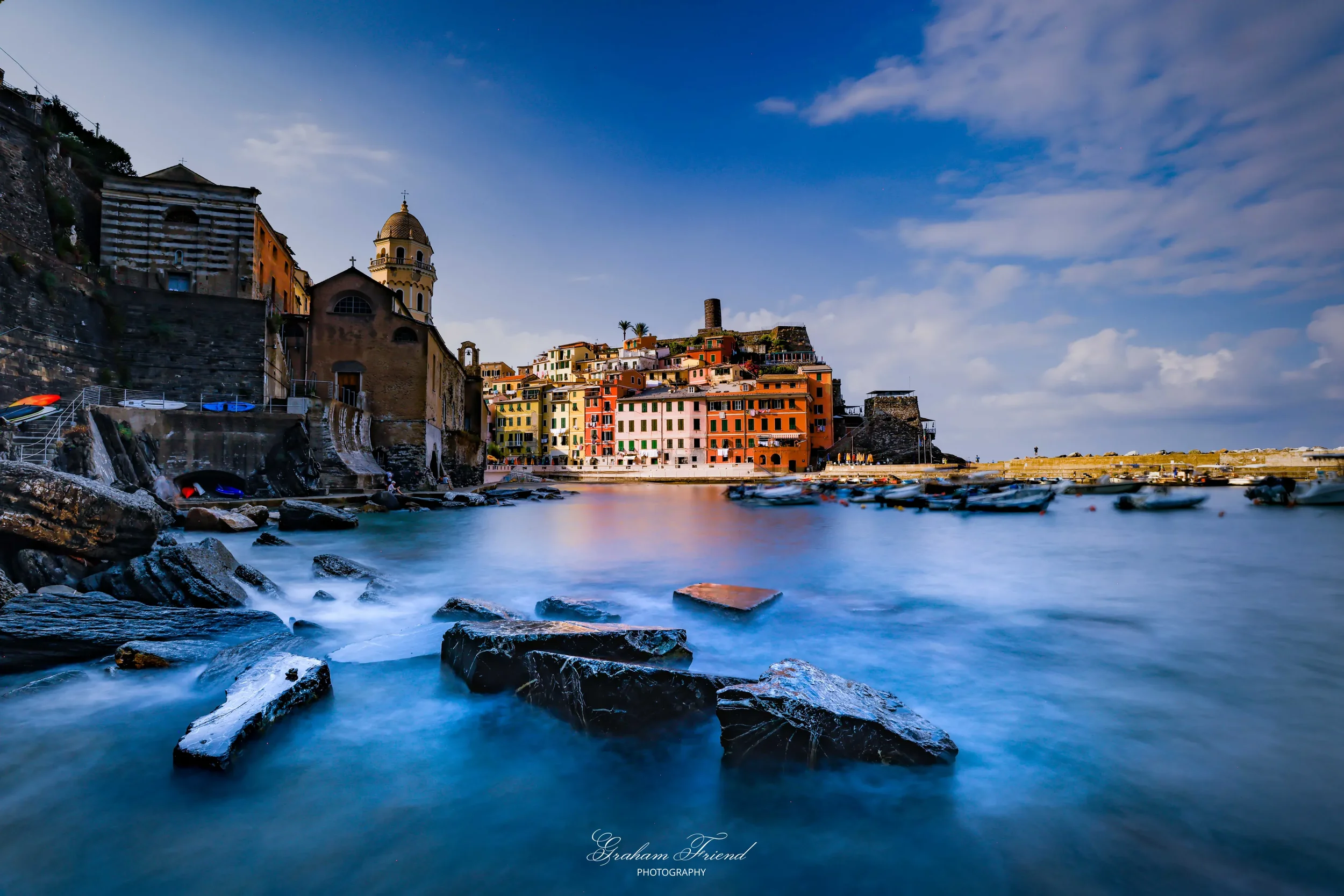 Colorful buildings along a rocky harbor in a coastal town with boats and a cloudy sky.