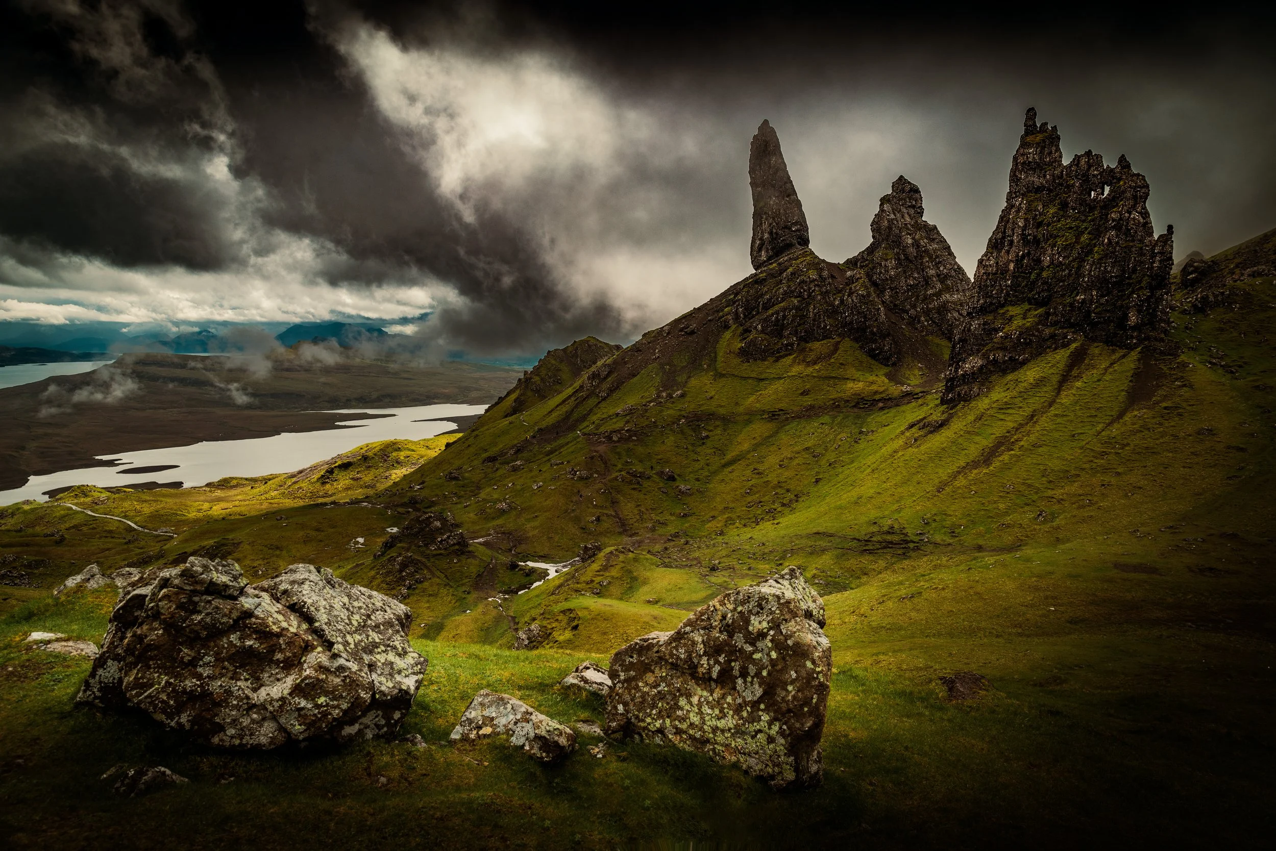 A dramatic landscape featuring green rolling hills, jagged rock formations, a lake, and dark storm clouds in the sky.