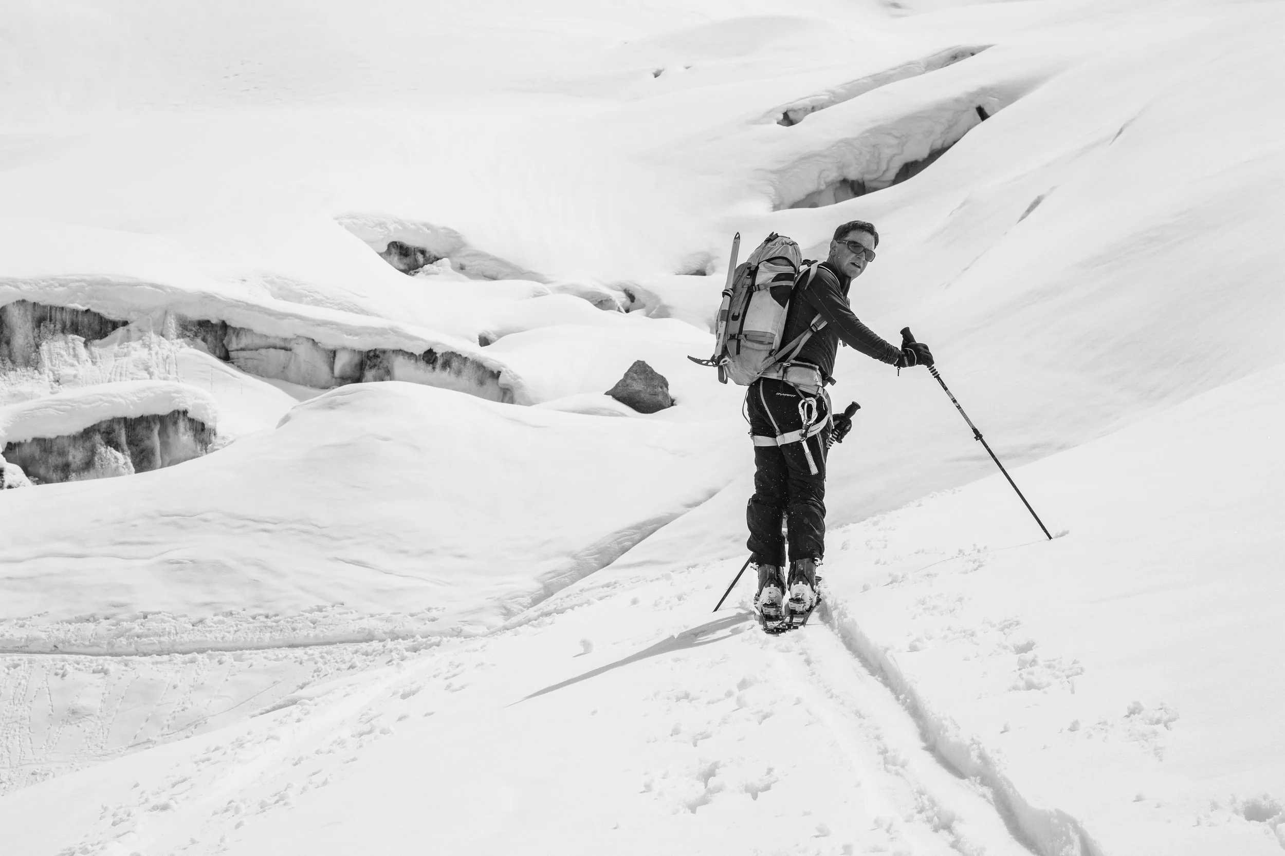 A person in winter gear with a backpack, using ski poles, crossing snow-covered terrain in a snowy mountain landscape.