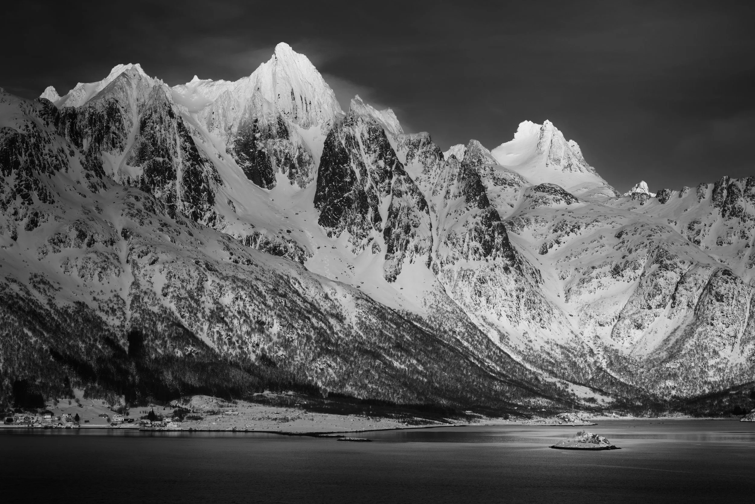 Black and white photograph of snow-covered mountains with a small house on an island in a calm lake in foreground.