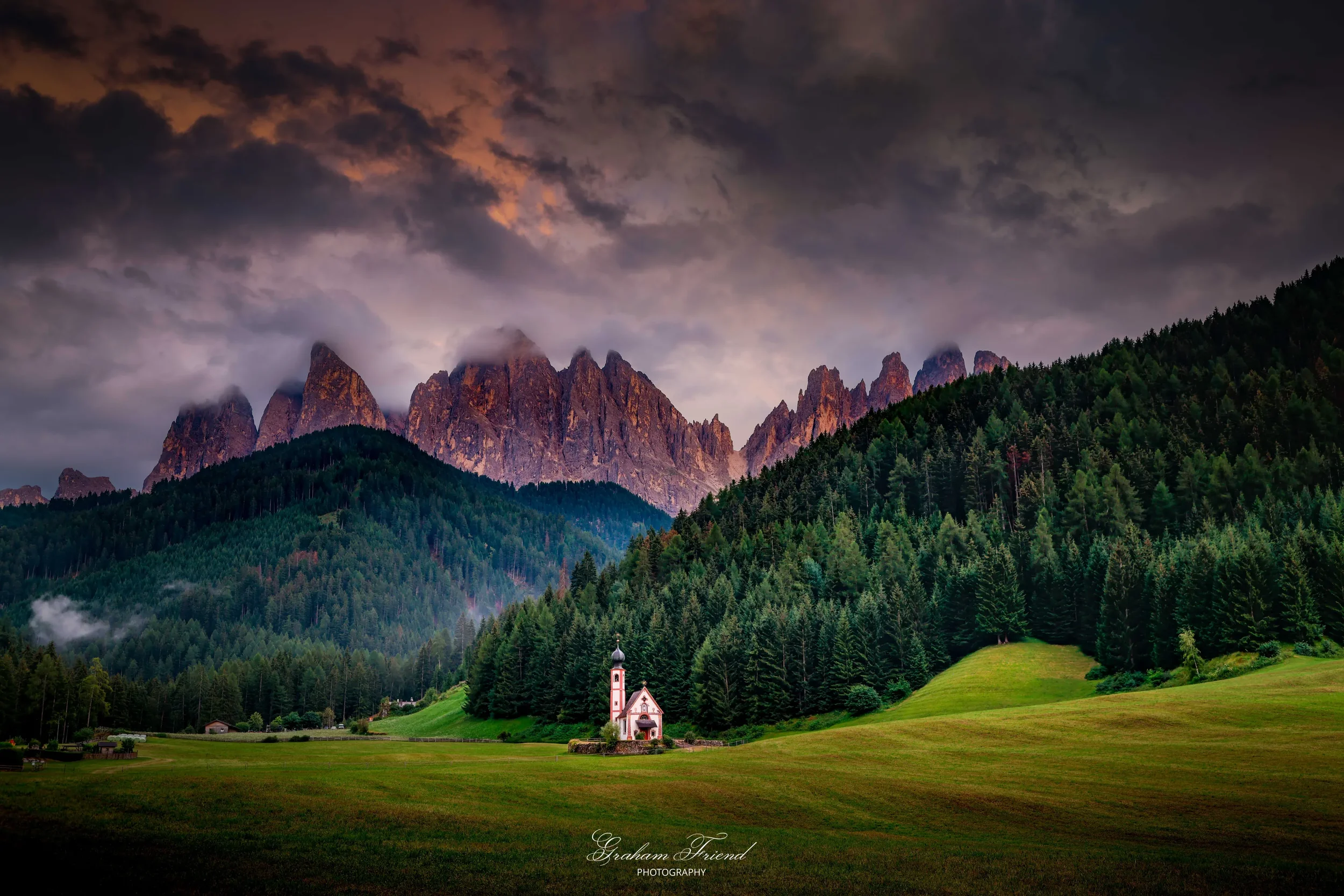 A small white church with a black steeple and cross is situated in a green meadow in front of a dense forest. In the background, rugged mountain peaks are partially obscured by clouds under a dramatic sky with orange and gray hues.