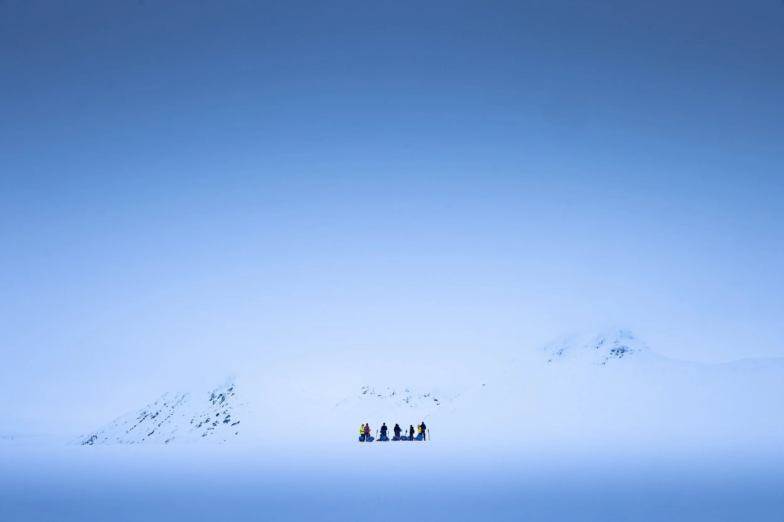 A group of people in winter clothing standing on snow in a vast, snowy landscape with snow-covered mountains in the background under a clear blue sky.