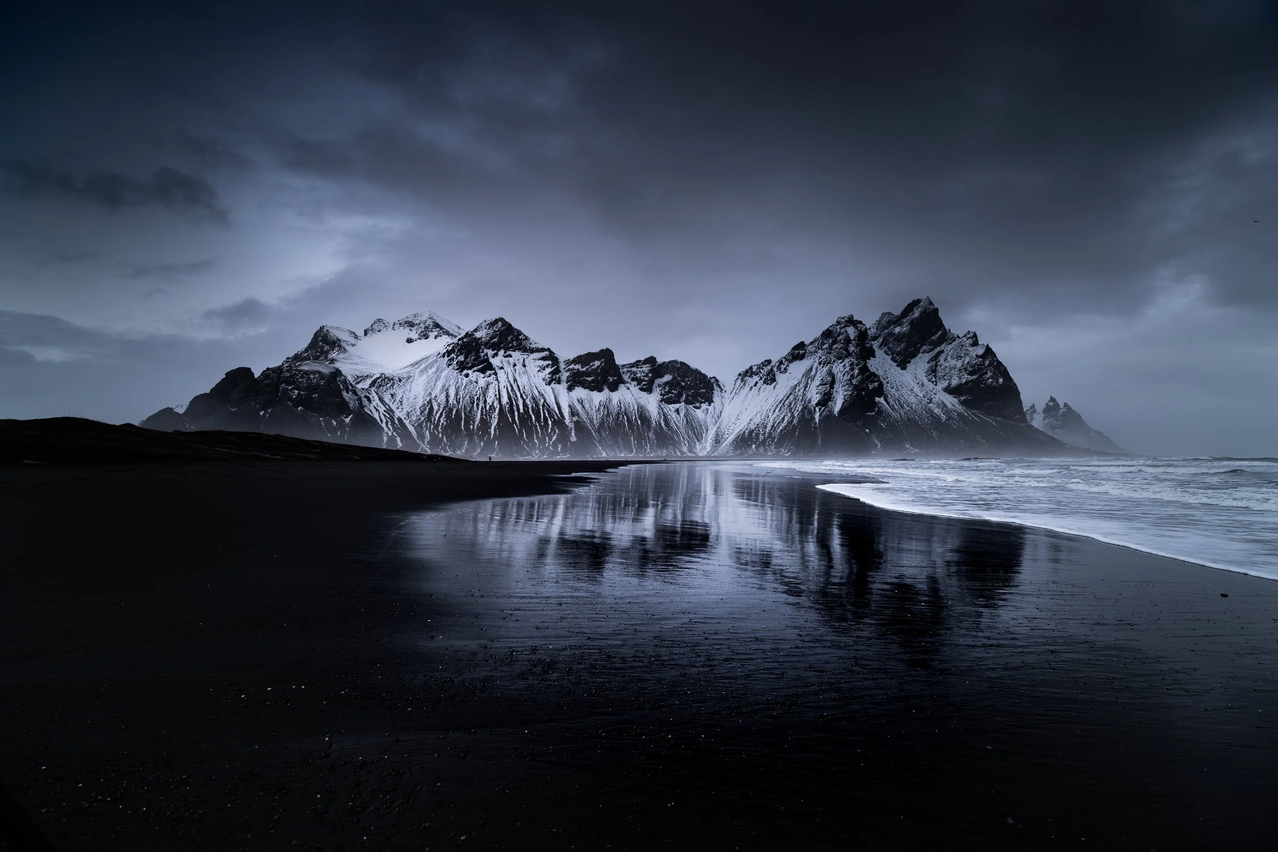 Dark, cloudy sky over snowy mountain range reflected in black sand beach and ocean waves.