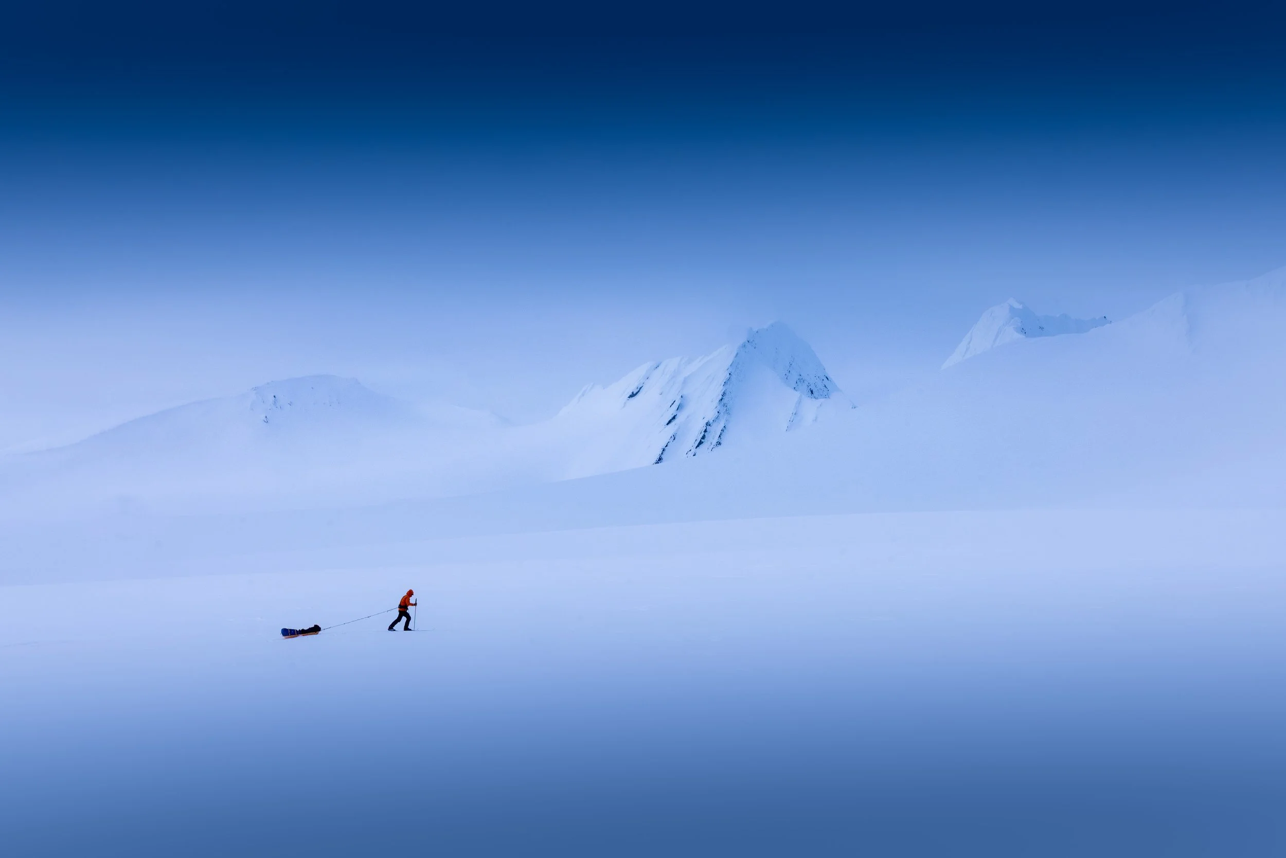 A person in an orange jacket pulling a sled across a vast, snowy landscape with snow-covered mountains in the background under a clear blue sky.