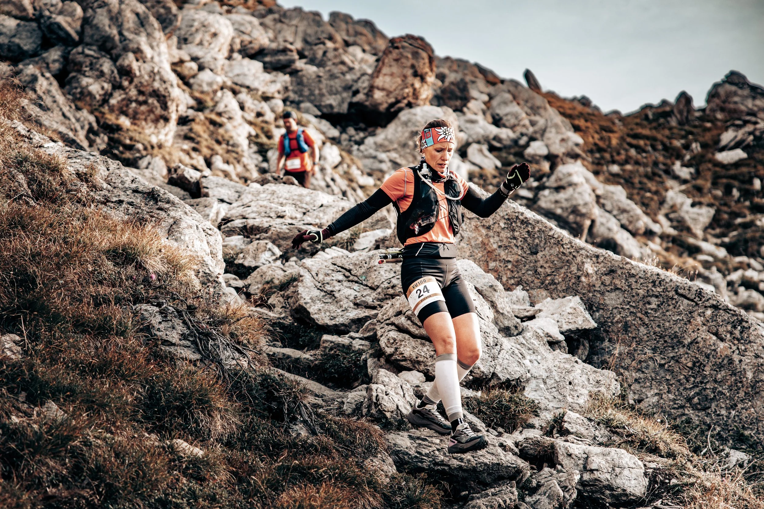 A female trail runner wearing a headband, black gloves, a black vest, orange shirt, black shorts with a race bib, and knee-high compression socks descending a rocky mountain slope.